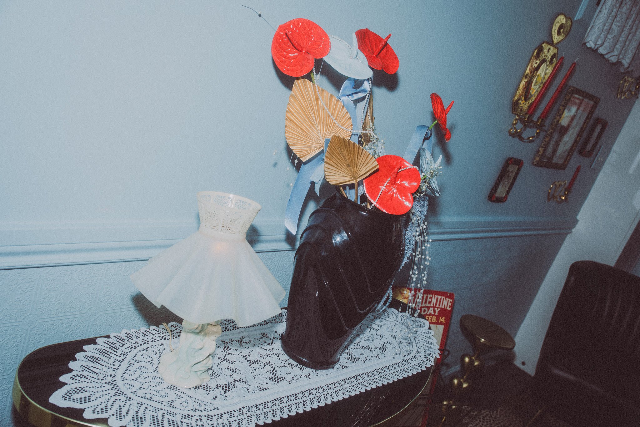 A decorated wall with paper fans and flowers in a black vase, a lace tablecloth with a small white lampshade on a wooden table, and a Valentine's Day sign in the background.