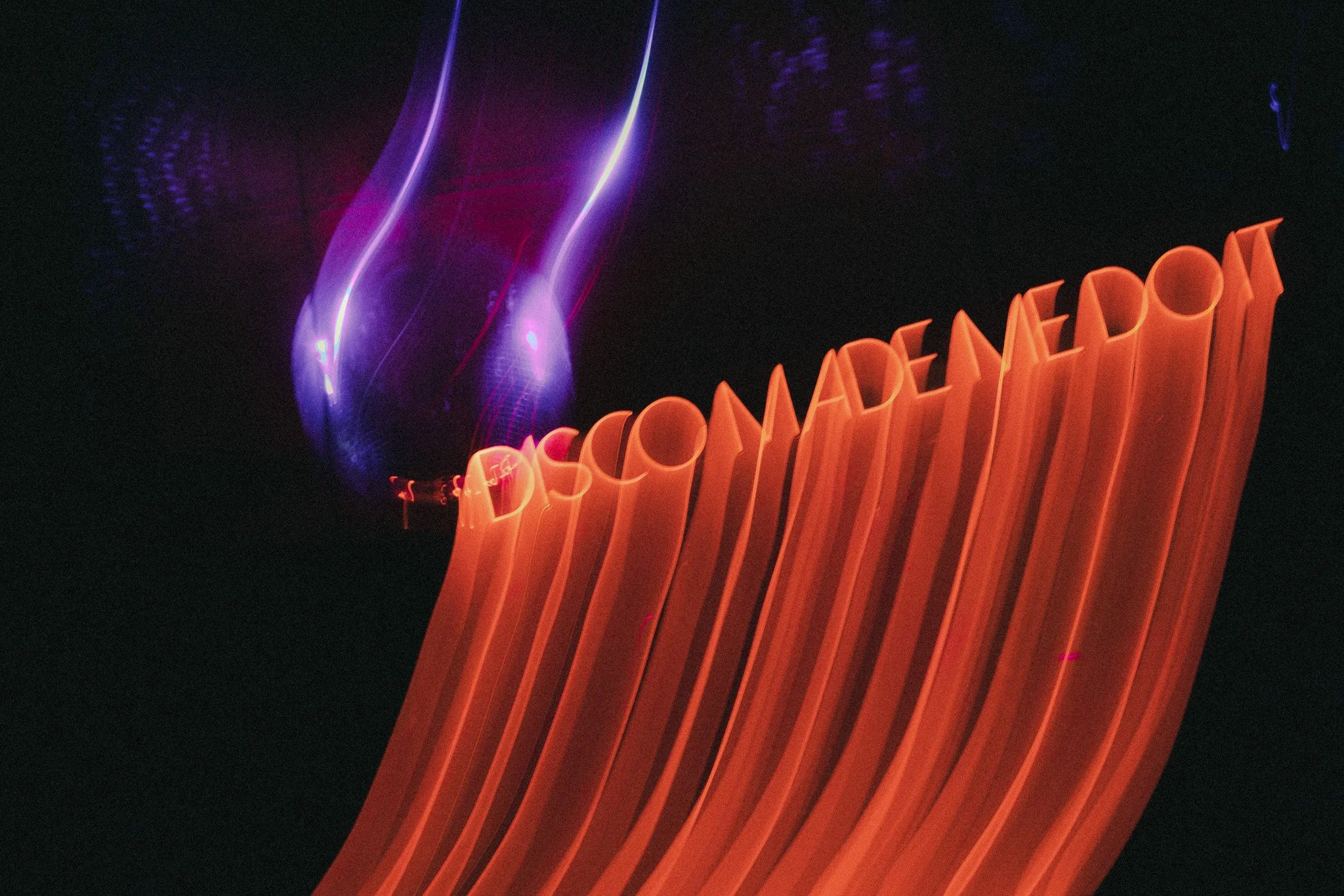 Neon sign displaying the message 'Discover About' in bright, glowing orange text against a dark background with violet light streaks.