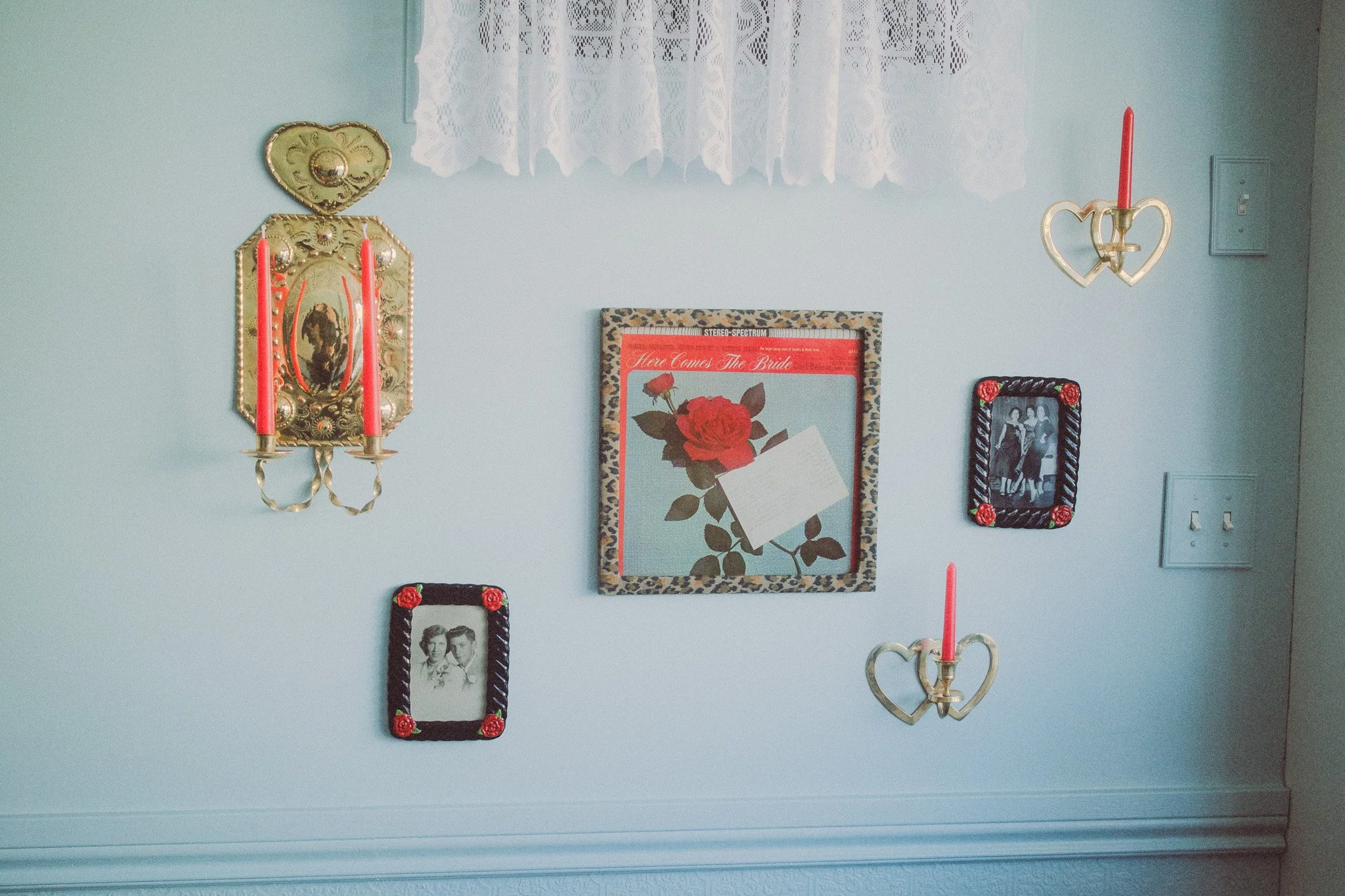 Wall decorated with vintage picture frames, heart-shaped candle holders, and floral artwork, with lace curtains at the top.