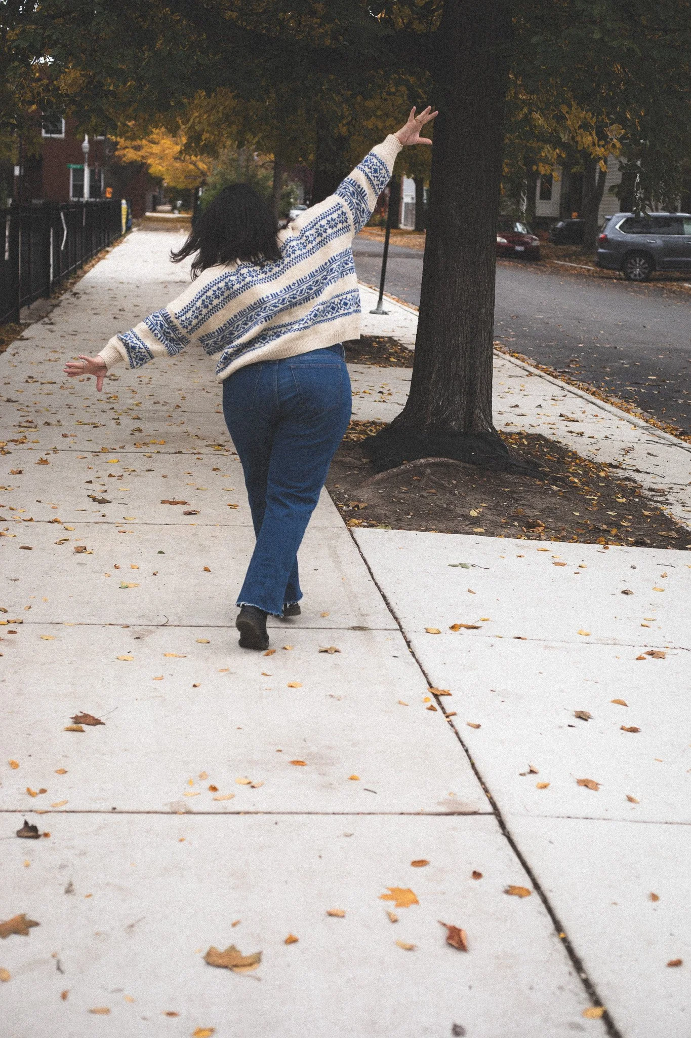 A person walking on a sidewalk in autumn, with trees and parked cars in the background, wearing a cream sweater with blue patterns and blue jeans.