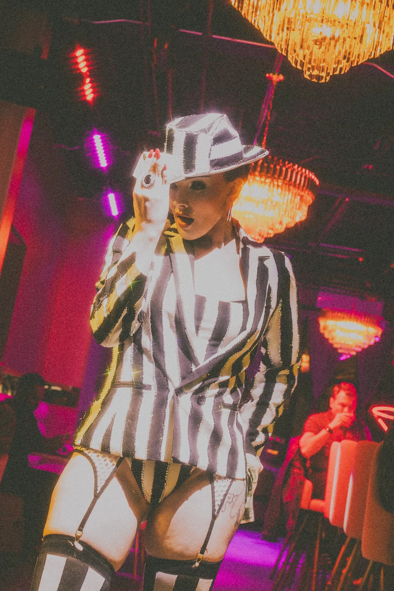 Person in a black and white striped outfit posing in a nightclub with colorful neon lights and chandeliers.