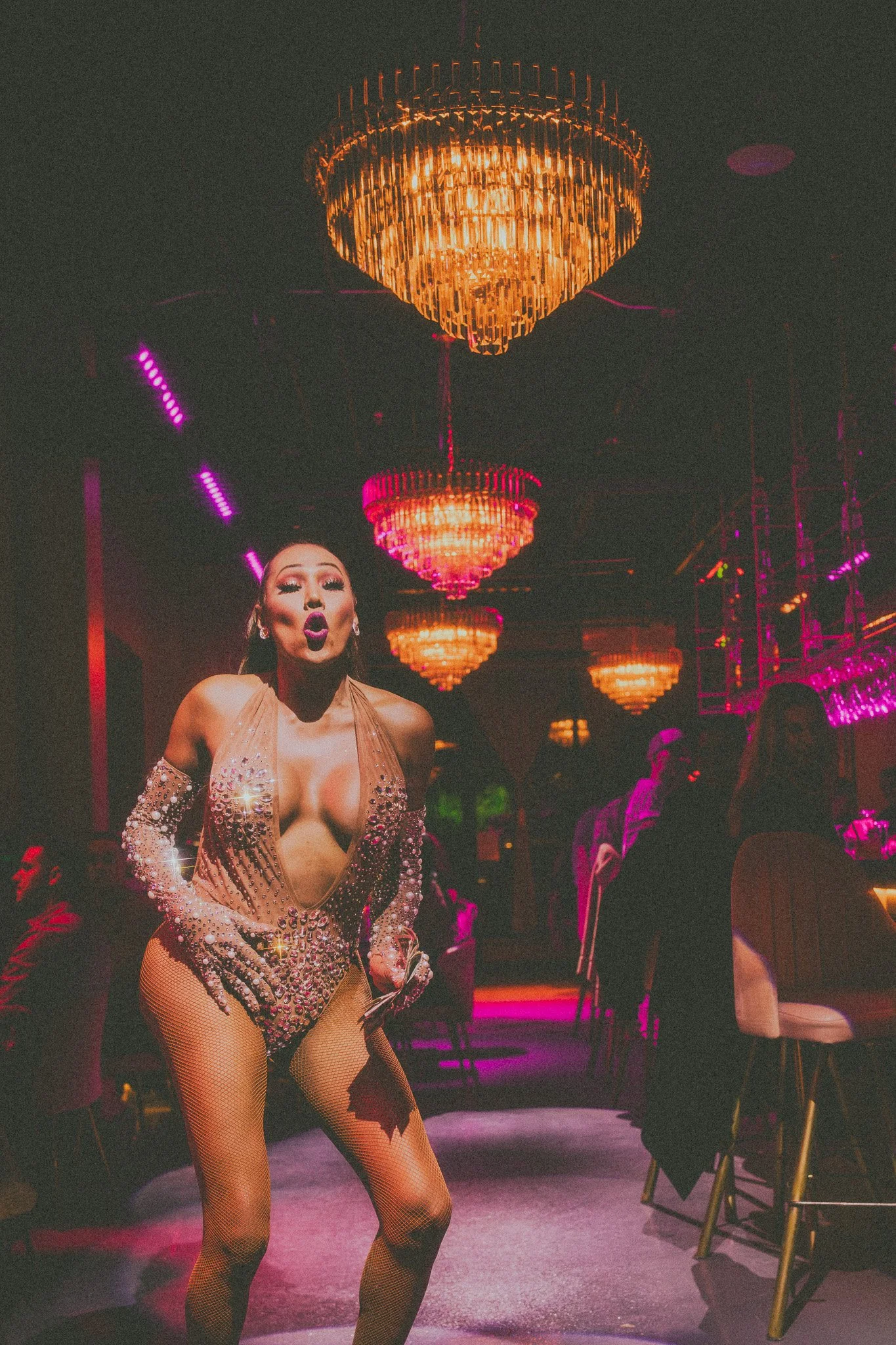 A performer in a sparkling, revealing costume dances in a nightclub with gold and pink chandeliers and colorful lighting.