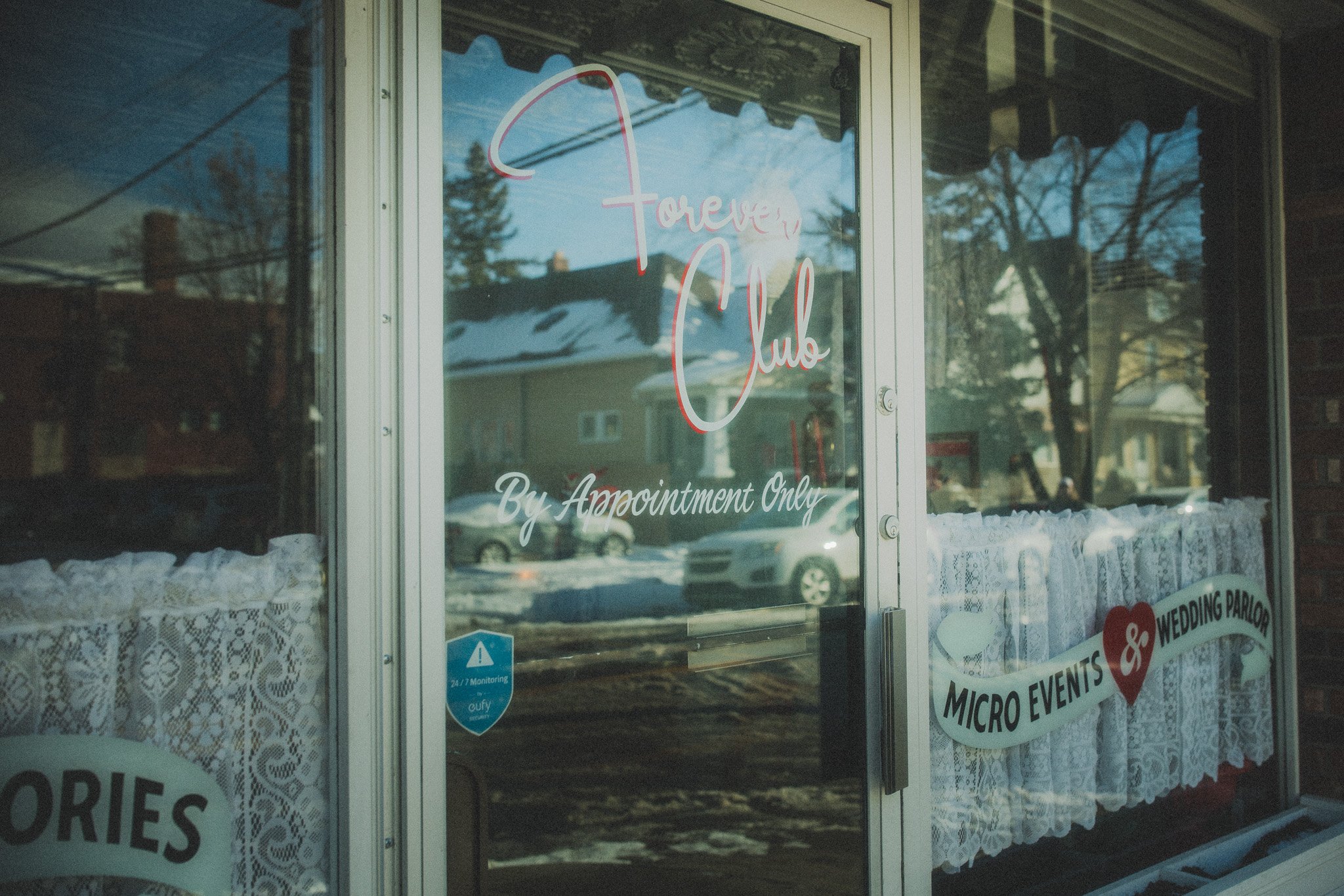 Glass door with the words "Forever Club" and "By Appointment Only" written on it. The door has lace curtains and signs indicating micro-events and wedding parlor. Reflection of cars and houses can be seen in the glass, with a snowy ground outside.