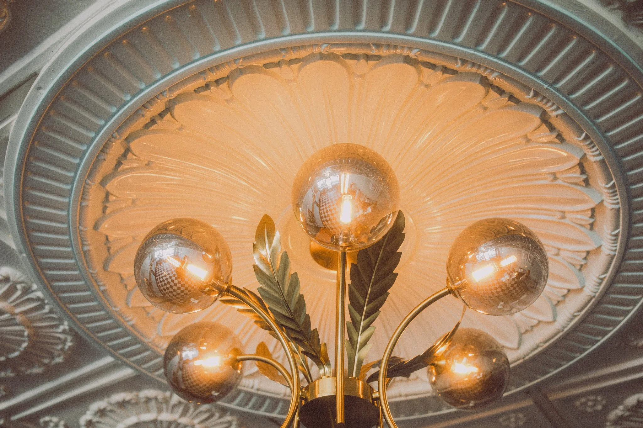 Close-up of a chandelier with multiple bulbs and decorative leaves, hanging from an ornate ceiling with detailed molding and a large central decorative element.
