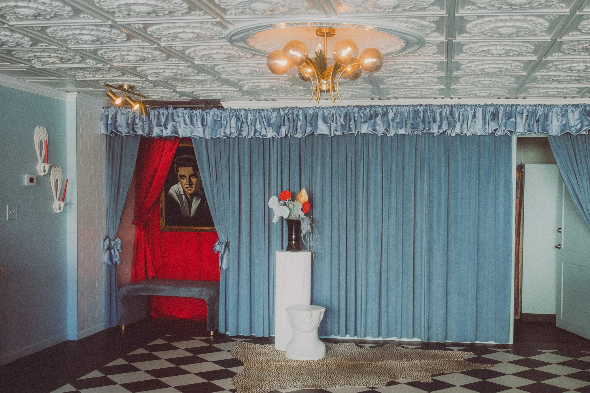 Room with blue curtains, a chandelier, a portrait of Elvis Presley on a red background, and a white pedestal with a black pitcher holding red, white, and orange flowers.
