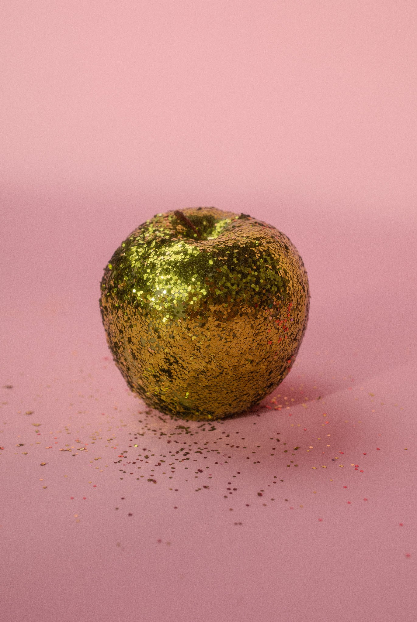 A golden apple covered in glitter sits on a pink surface with scattered glitter around it, against a pink background.