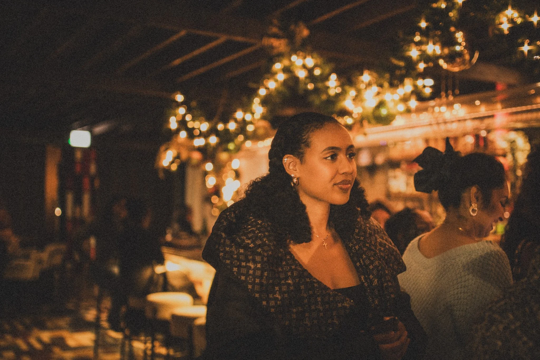 Woman with curly hair and earrings at a dimly lit celebration or dinner event, decorated with string lights and ornaments in the background.