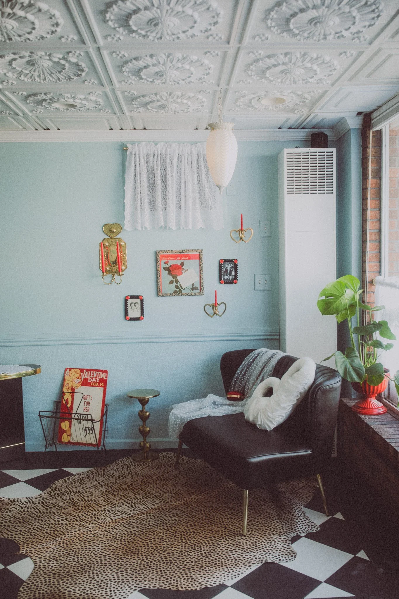 A cozy living room corner with a black leather sofa, white decorative pillow, and a white swan-shaped pillow. There's a small gold side table, a leopard print rug, and a window with lace curtains. The wall has vintage-style decorations and framed pho