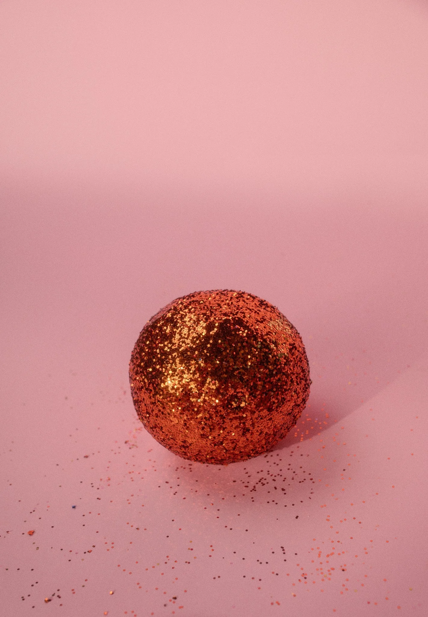 A glittery red-orange sphere on a pink surface with scattered glitter around it.