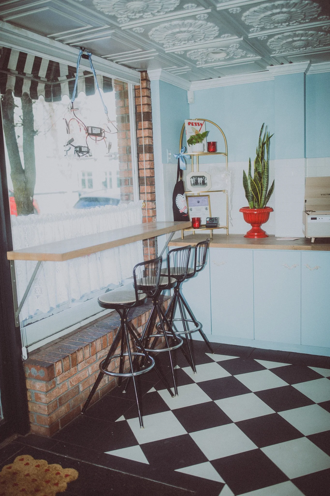 A cozy kitchen corner with black and white checkered floor tiles, a small bar-height counter with three black metal chairs, a large window with curtains, a blue wall with white cabinets, a plant in a red pot, small decorative items, and a ceiling wit