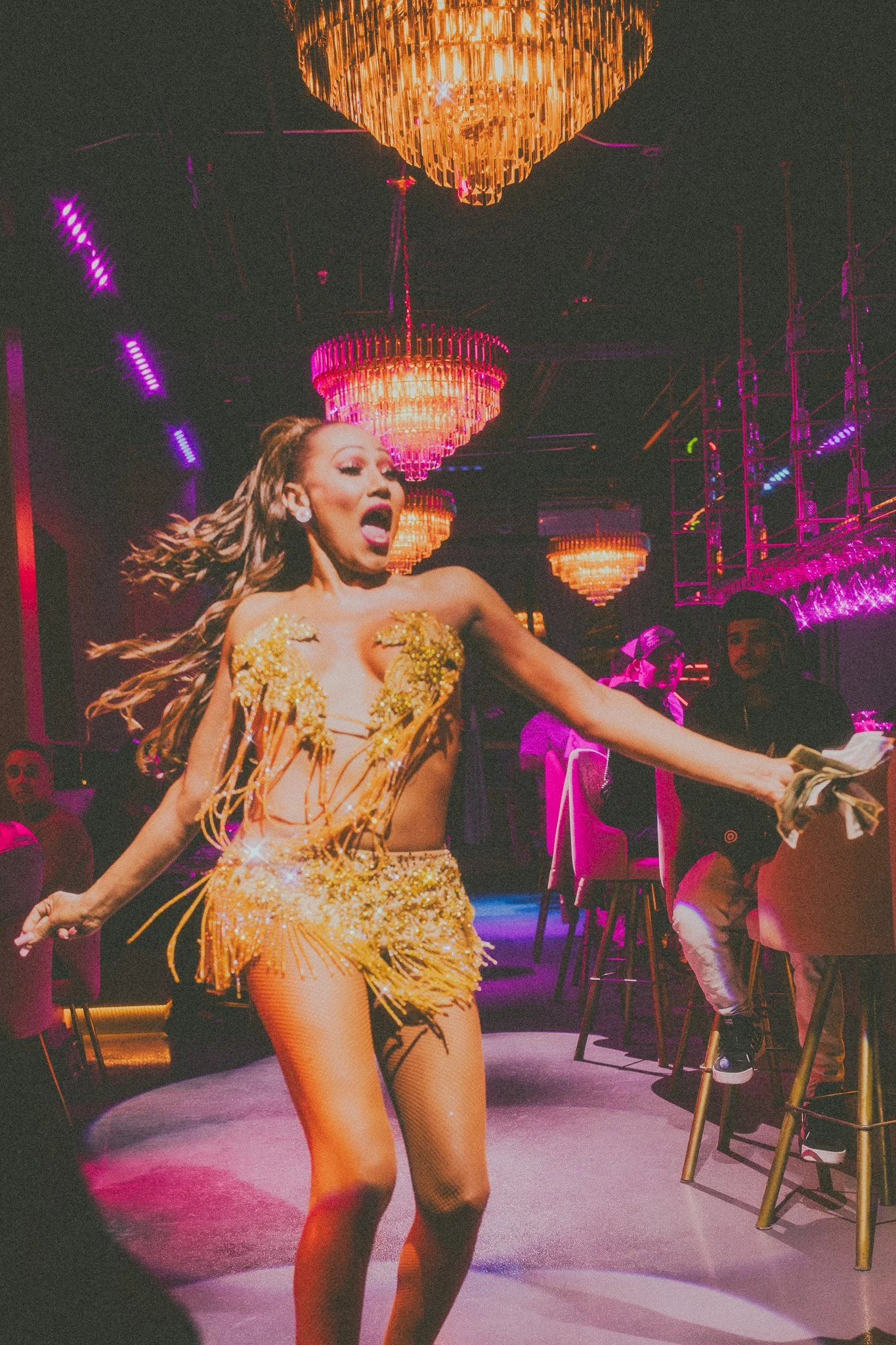 A woman performing a dance moves on stage in a glamorous gold outfit with fringe, under colorful chandeliers in a nightclub setting.