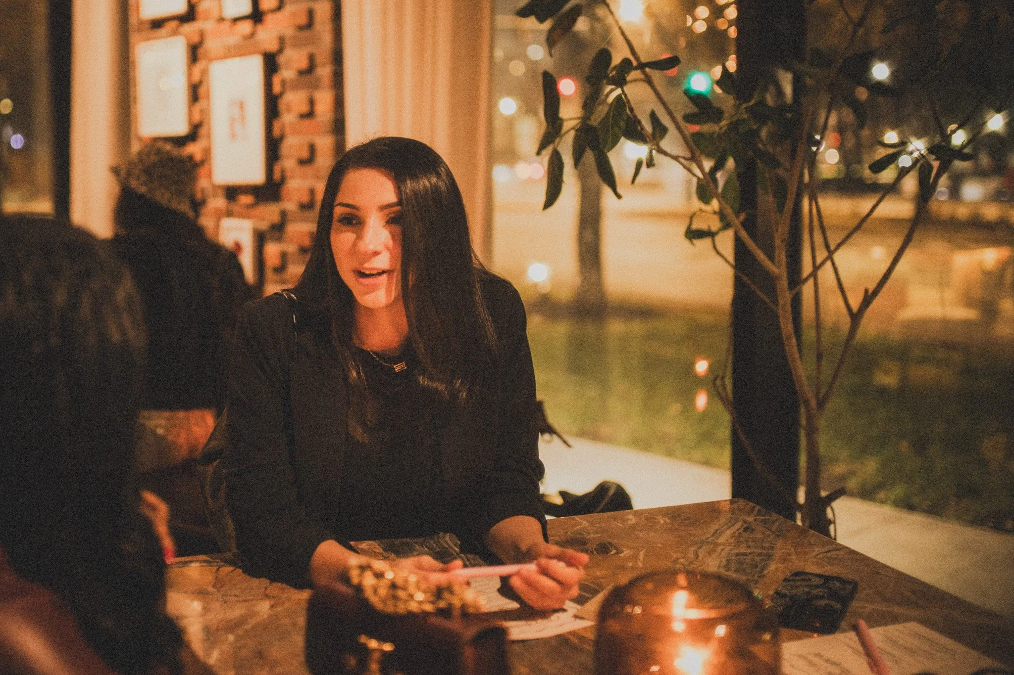 A woman with long dark hair, sitting at a table in a cozy restaurant or cafe during the evening or night. She is holding a pen and appears to be talking or smiling, with a candle, phone, and some papers in front of her. The background features warm l