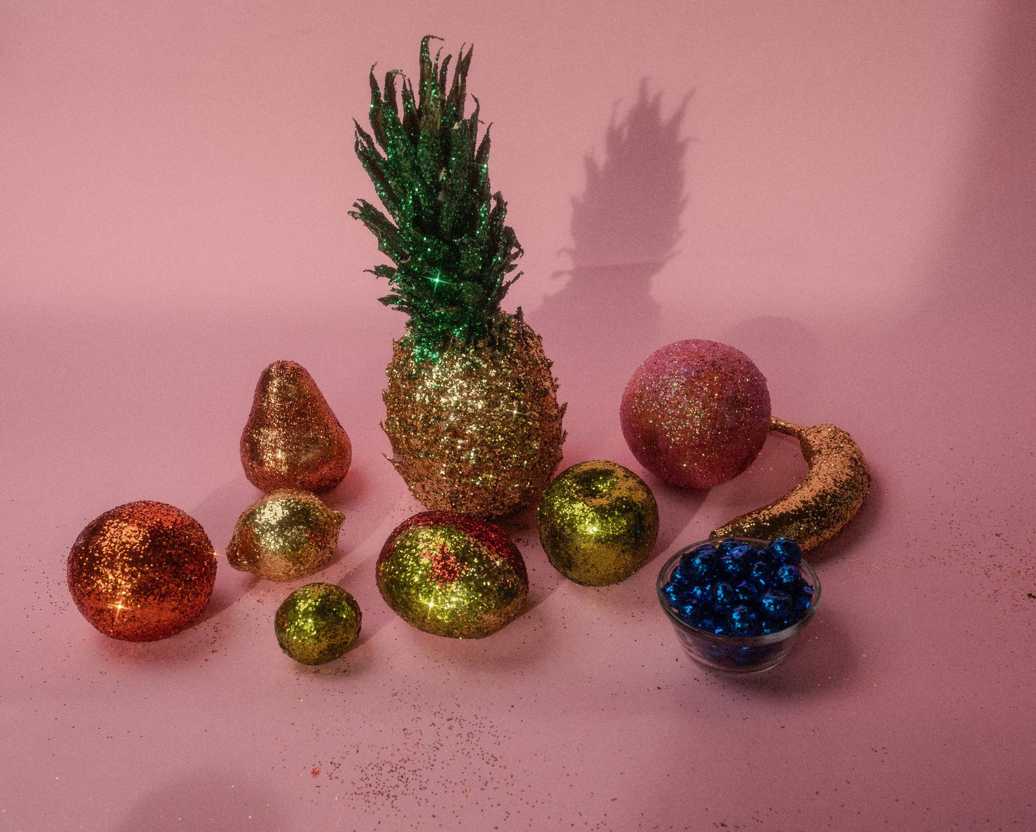 Decorative glittery fruits and a small bowl of blueberries on a pink surface with a pink background.