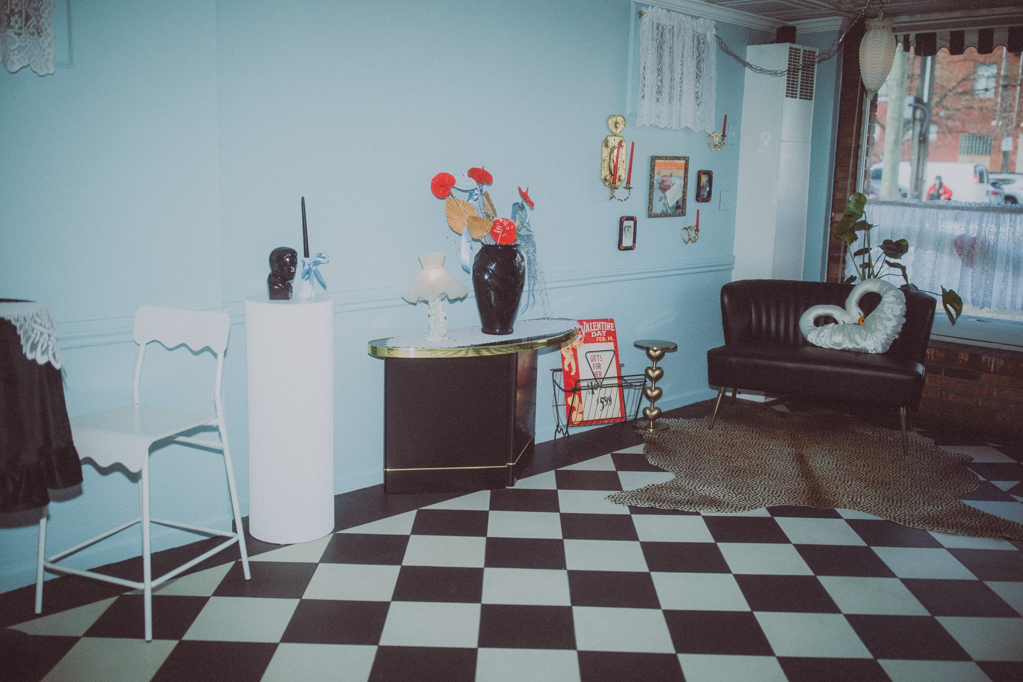 Vintage-style living room with black and white checkered floor, black leather sofa with swan-shaped pillow, small round table with plant, and wall decor including framed pictures and red candle holders.