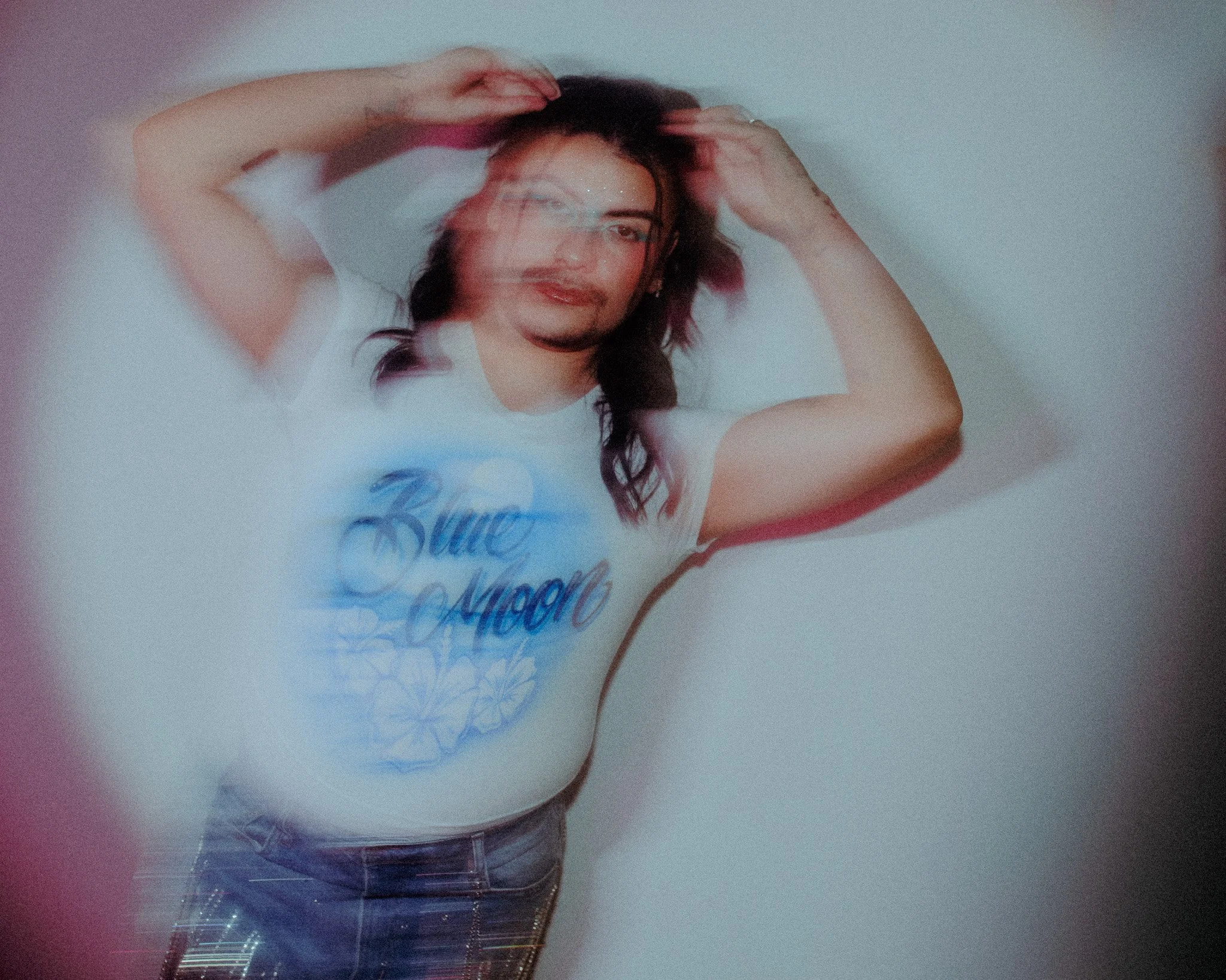 A person with long dark hair, holding their hands to their head, lying on a white surface, wearing a white t-shirt with blue and purple text.