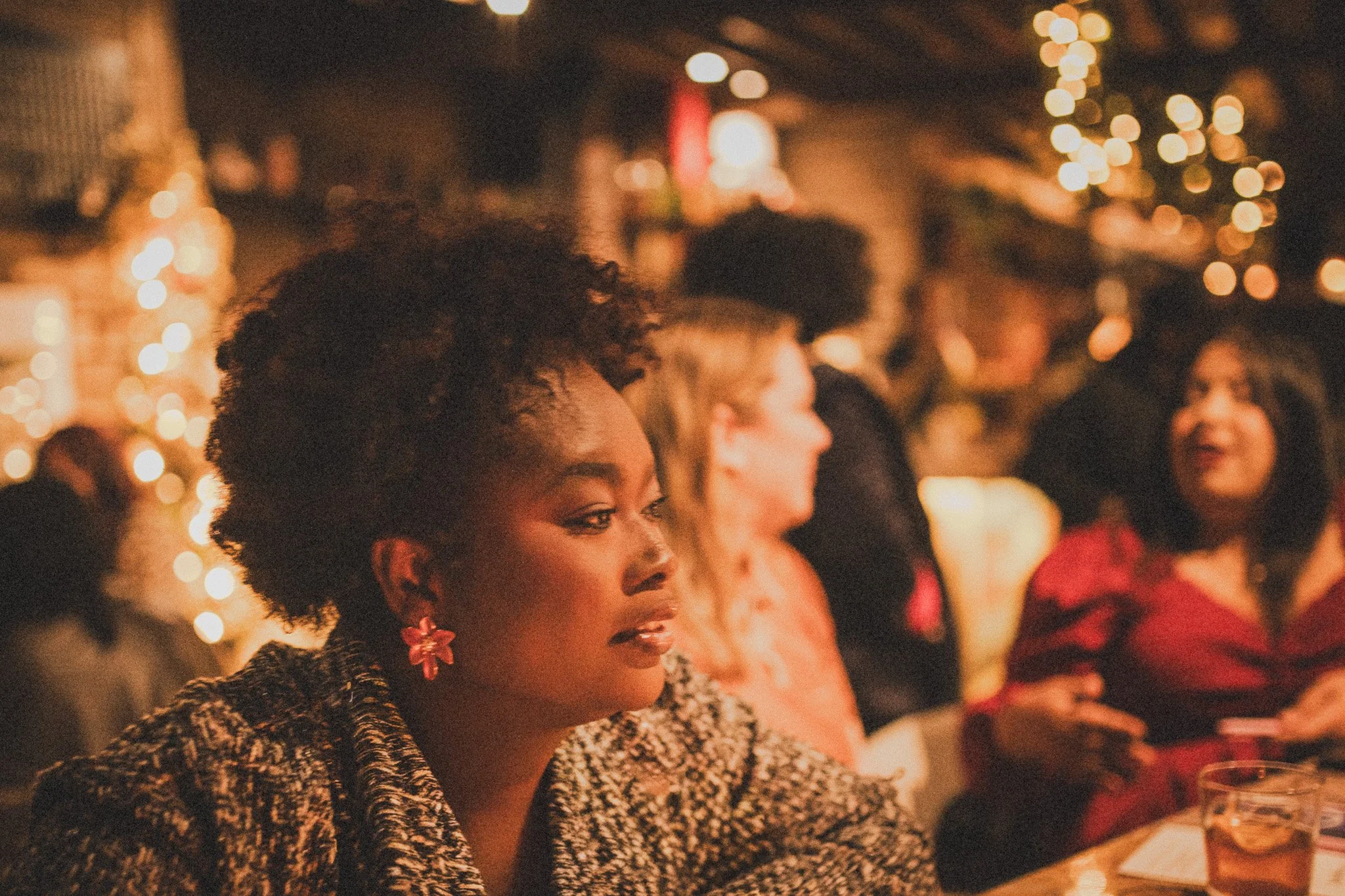 A woman with short, curly hair and pink flower earrings sits at a table in a warmly lit, festive setting with blurred string lights, with other women around her, some in red attire.