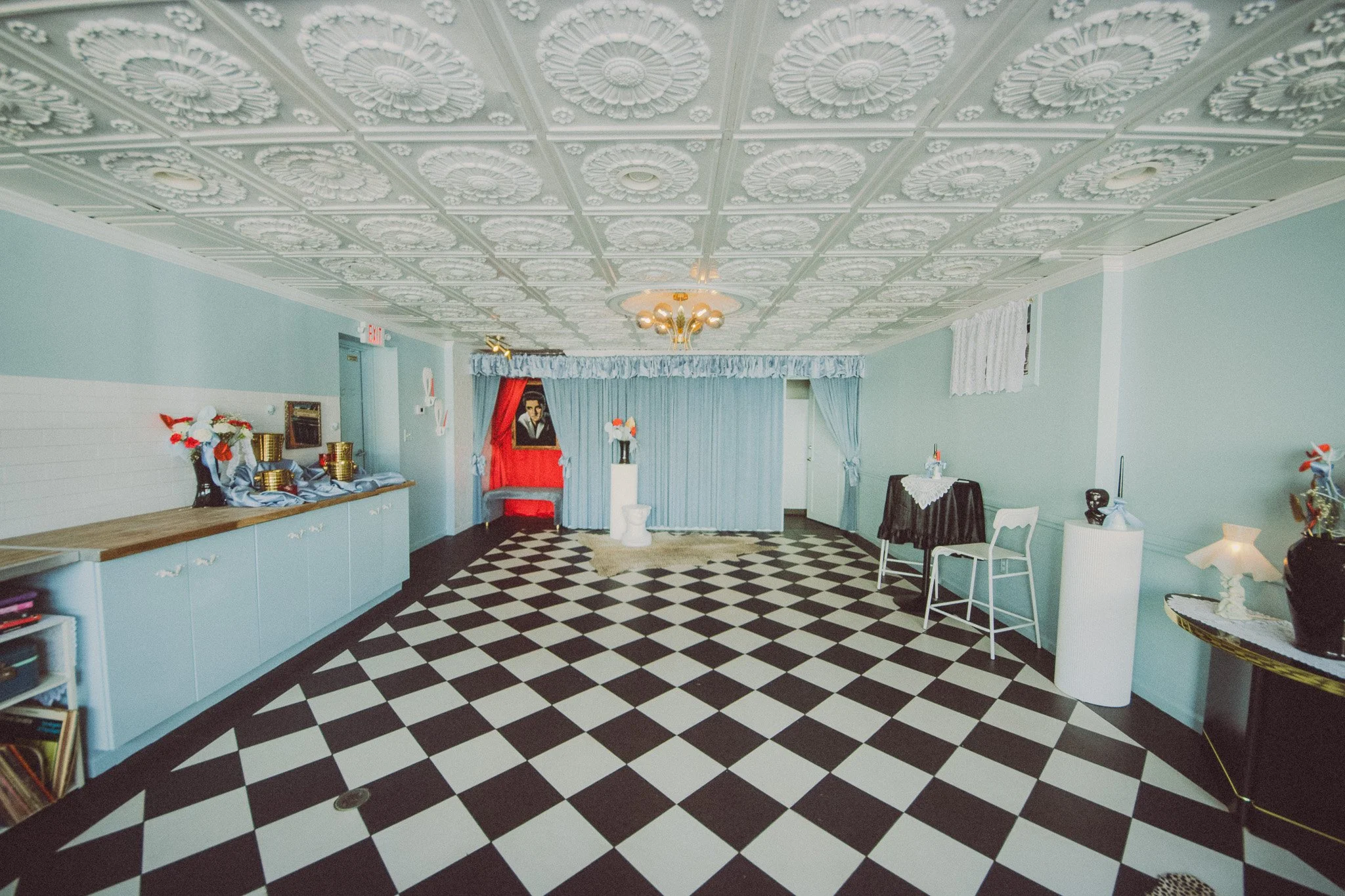 Decorated interior with checkered black and white floor, light blue walls, vintage ceiling tiles, small tables with items, and a puppet theater resembling Elvis Presley.