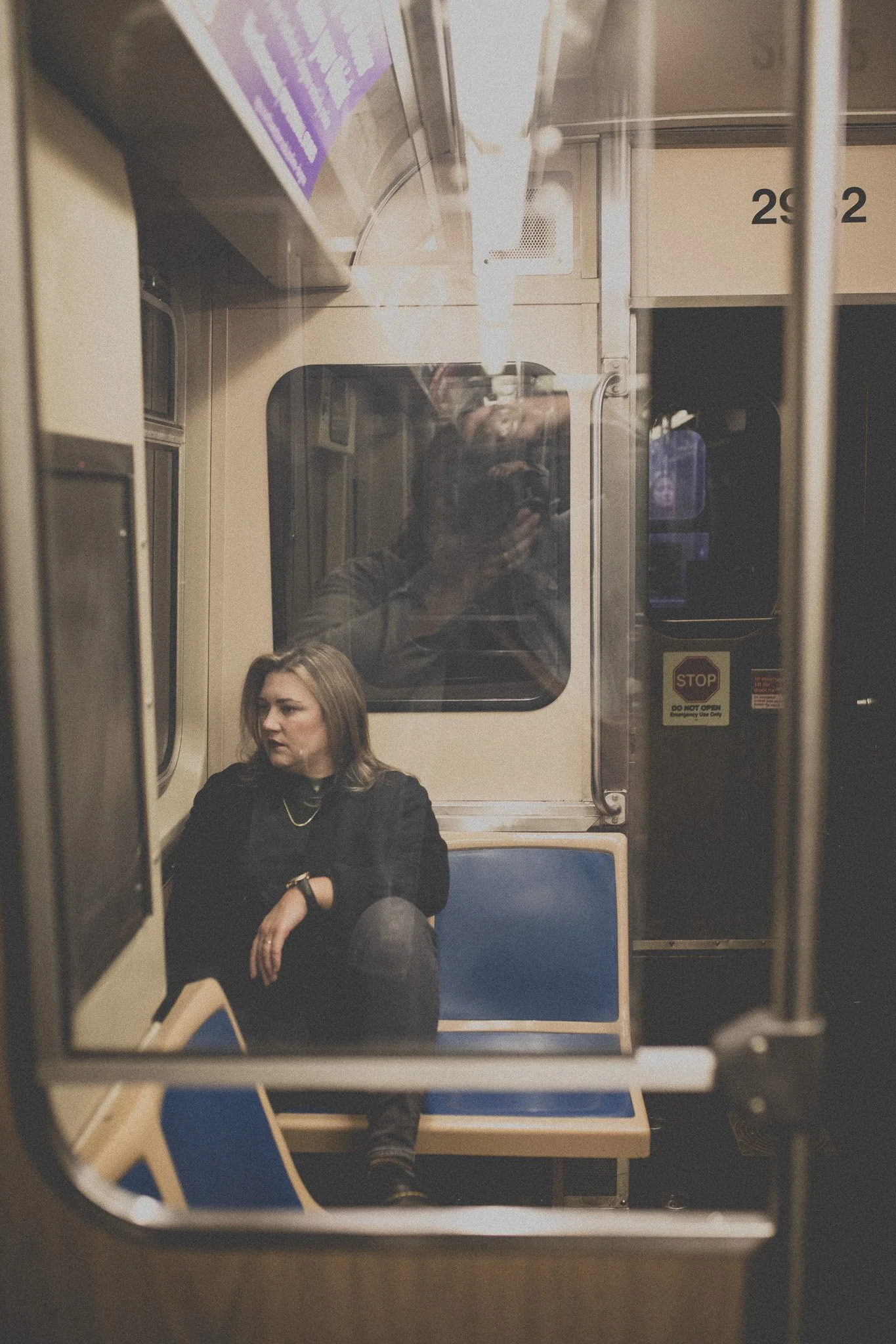 A woman sitting alone on a train looking to the side, with her arms crossed, and a reflection of a person taking the photo visible in the window behind her.