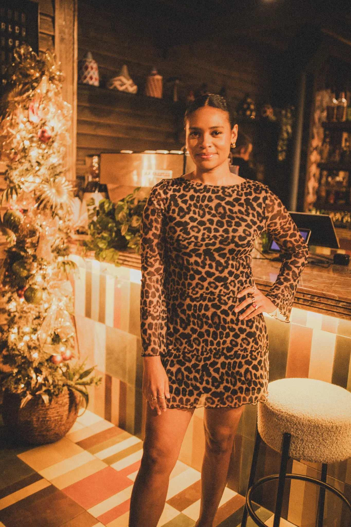 A woman in a leopard print dress standing next to a decorated Christmas tree in a warmly lit indoor setting.