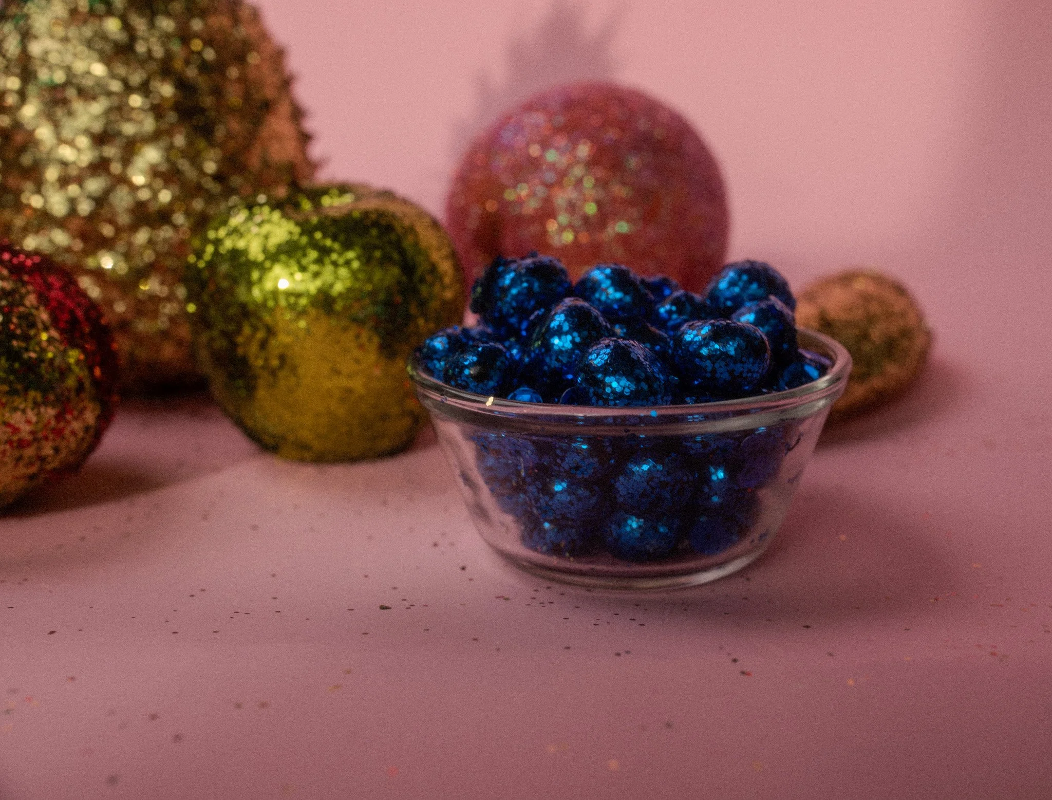 Blue glittered decorative objects in a glass bowl with gold and pink glittered ornaments in the background on a pink surface.
