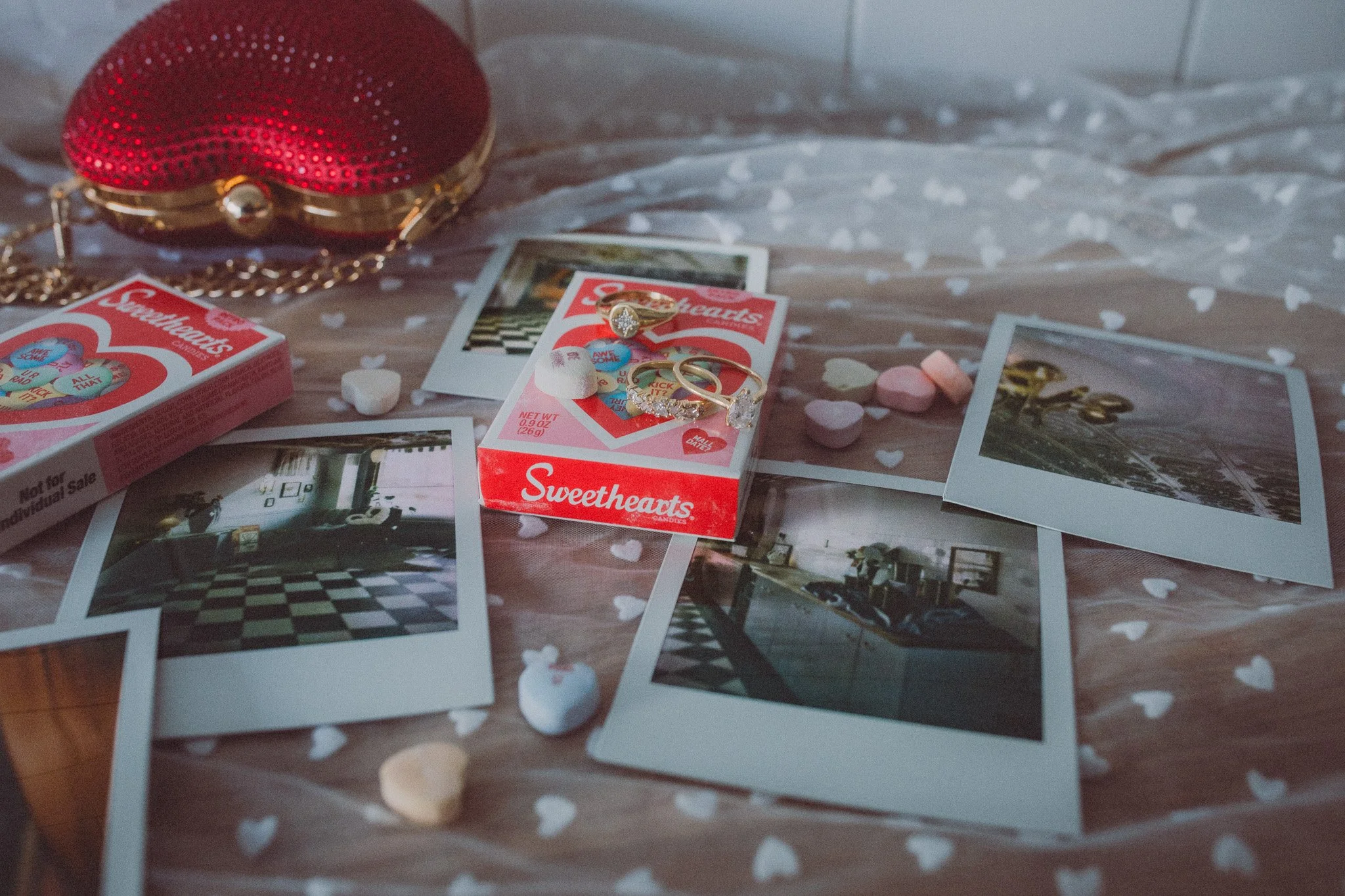 Polaroid photos, a red Sweethearts candy box, a heart-shaped purse, rings, and heart-shaped confetti on a heart-patterned surface.