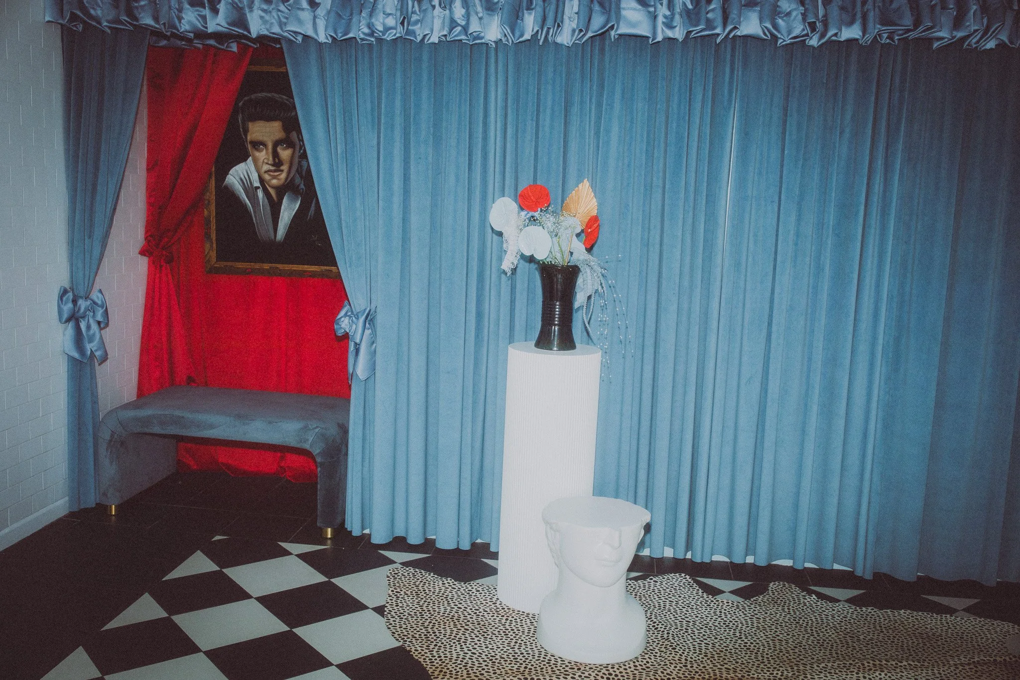 A room with blue curtains, a portrait of a woman on the wall, a bench covered with a gray cloth, a black vase with red, white, and beige paper flowers on a white pedestal, and a white bust-shaped stool, with a black and white checkered floor and a be