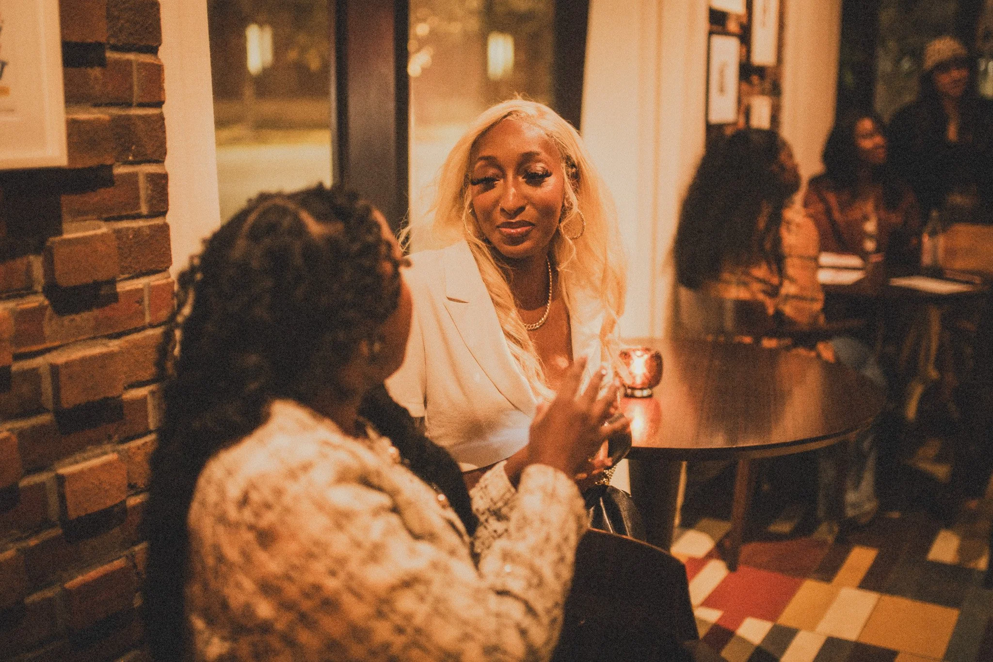 Two women sitting at a table in a warmly lit indoor setting, engaged in conversation. The woman on the right has long blonde hair, is wearing a white blazer, and is holding a glass. The other woman has dark curly hair and is wearing a patterned outfi