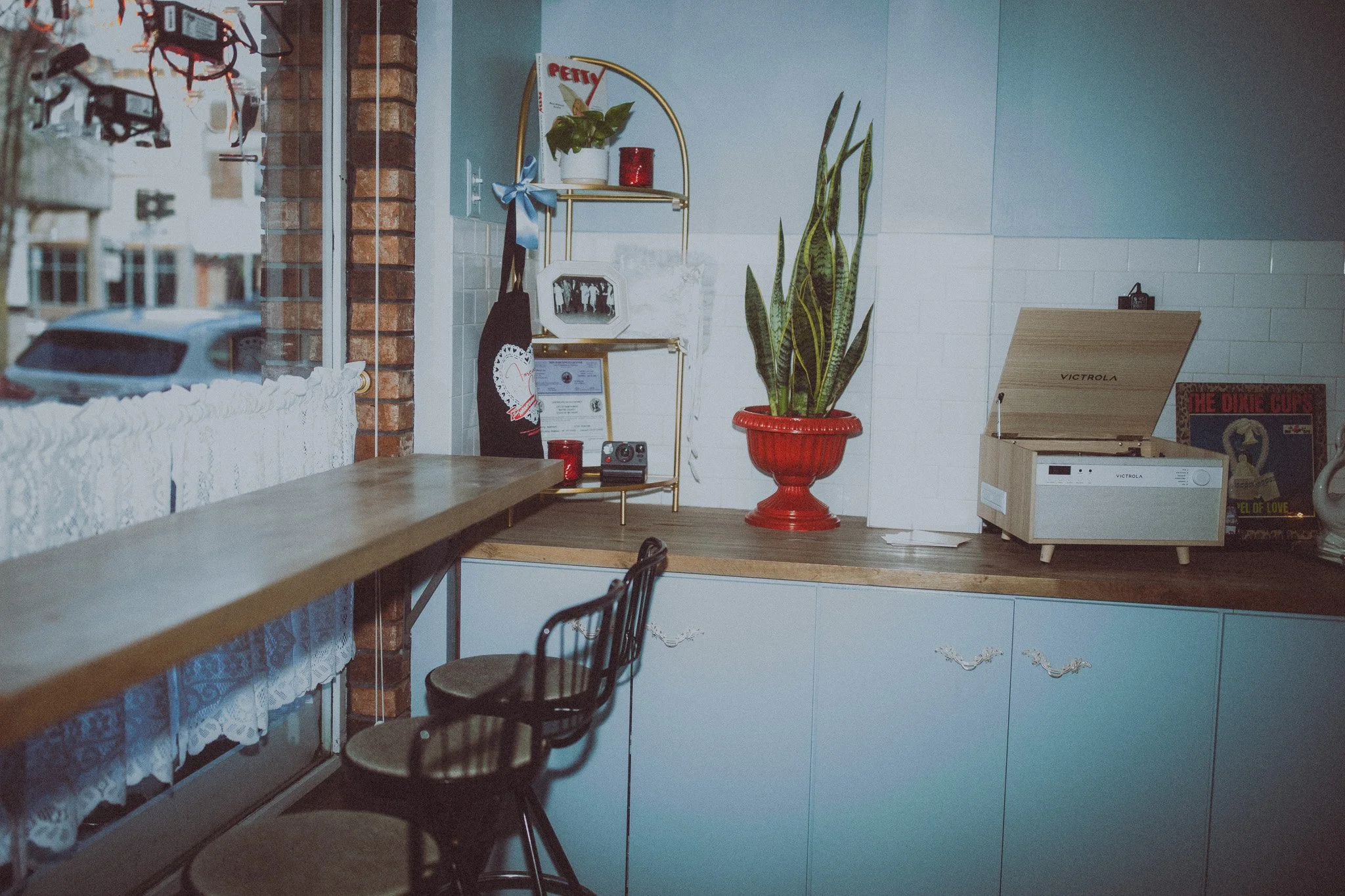 A cozy kitchen interior with a wooden countertop, a large potted snake plant in a red planter, a vintage record player, a framed picture, some framed documents, and a decorative shelf with a small plant and red candle, with a window showing a street 