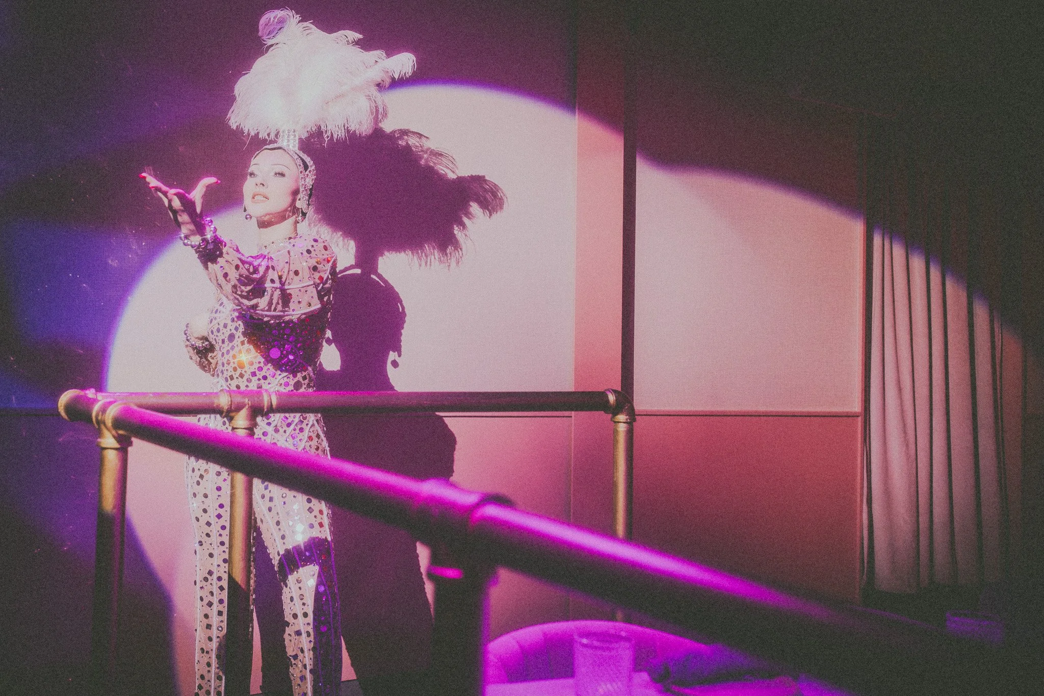 Person dressed in a glittery, polka-dotted costume with a large feathered headpiece performing in front of a colorful, illuminated background.