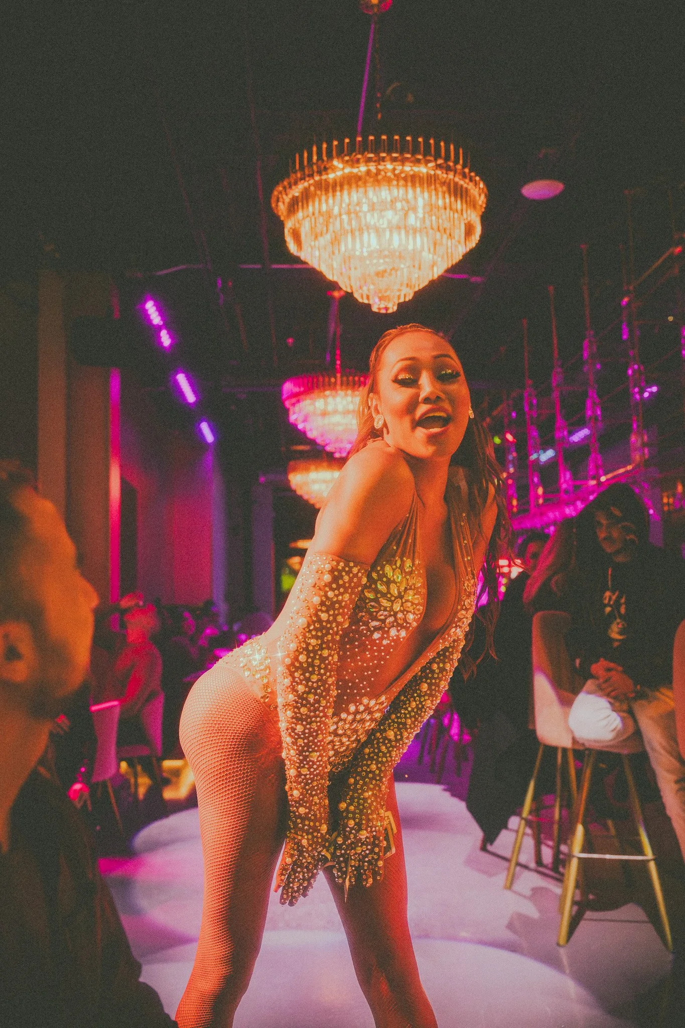 A woman performing on stage in a sparkly, revealing costume with rhinestones, in front of a chandelier and colorful lighting, at a nightclub or similar venue.