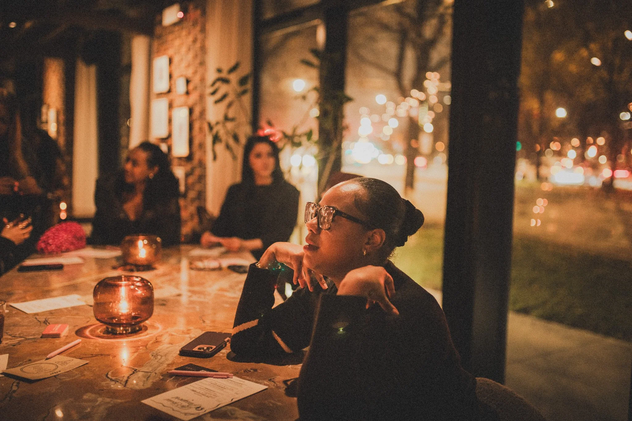 Group of women sitting at a wooden table in a cozy restaurant during evening, with warm lighting and city lights visible outside.