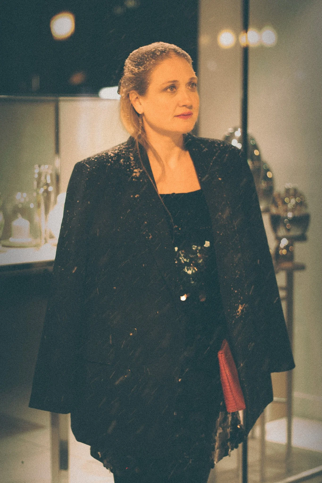 A woman with blonde hair in a black sequined blazer standing indoors at night with blurred lights in the background.