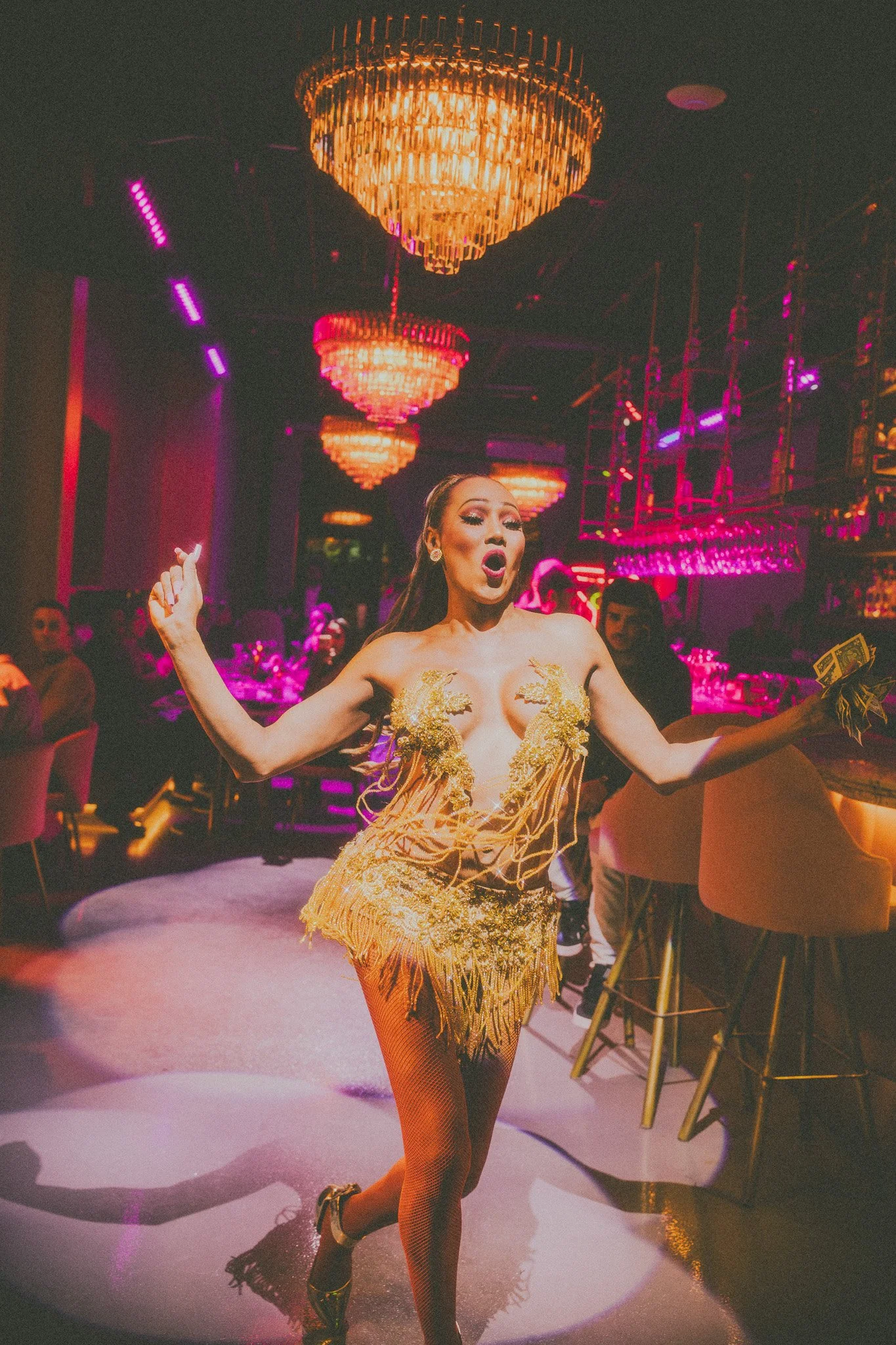 A performer in a gold, fringe costume with gold embellishments, singing or performing in a nightclub with pink and purple lighting and chandeliers overhead.