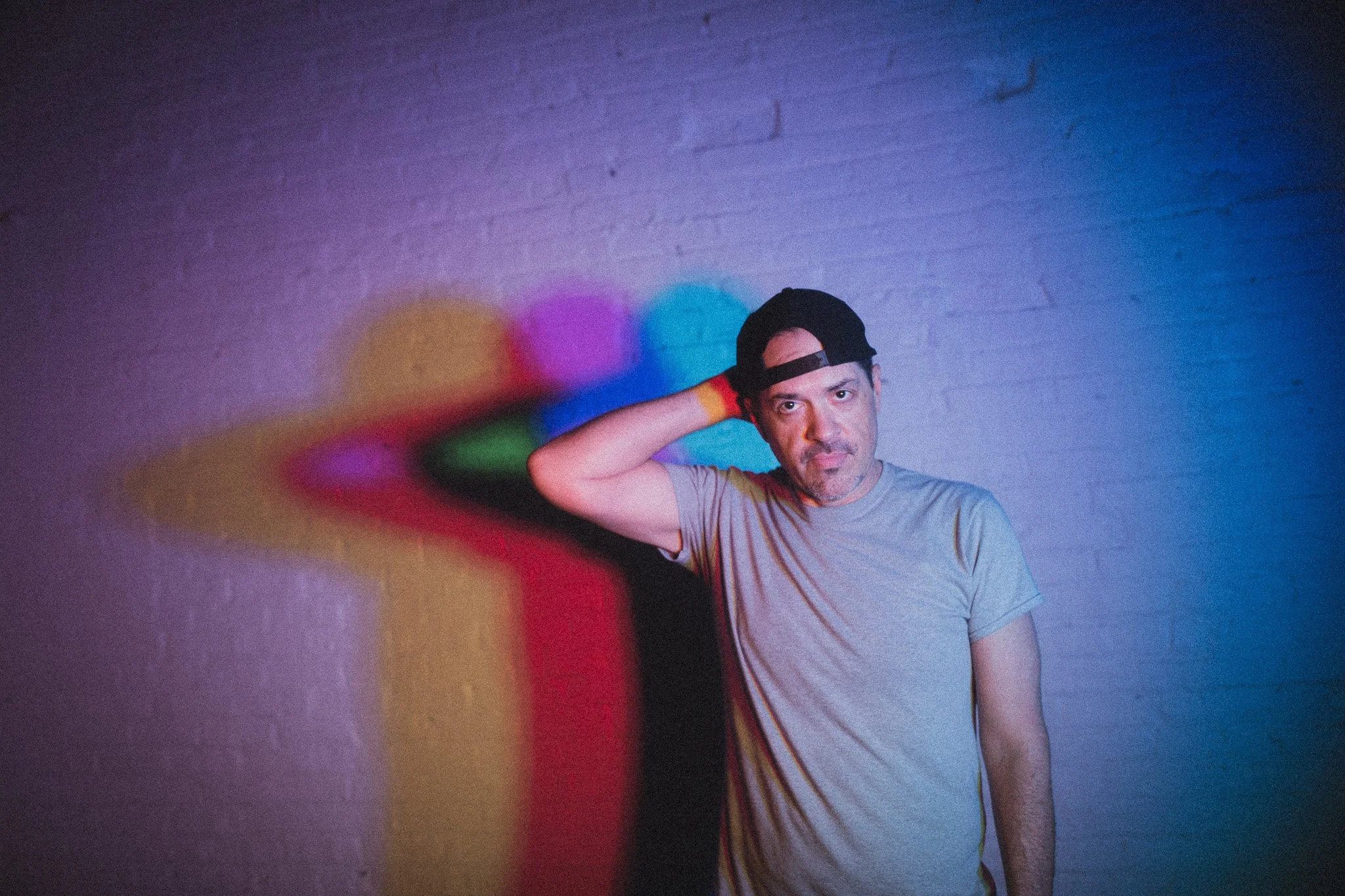 A man wearing a gray t-shirt and a black cap poses against a gray brick wall. He touches the back of his head with his right hand. Colorful, blurred circular lights are projected onto the wall behind him.