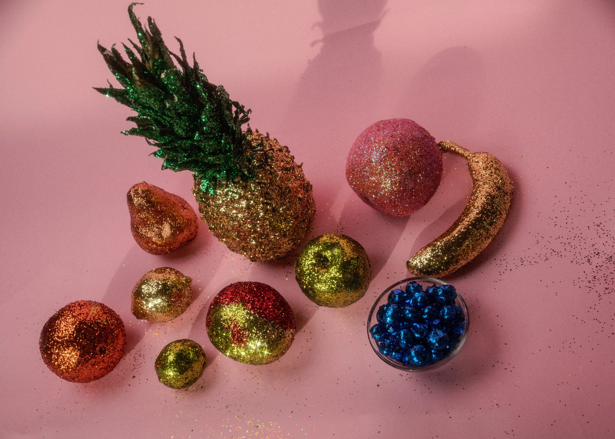Glittery, colorful holiday decorations including a pineapple, strawberries, a green and a red ball, a gold ball, a glittery banana, and a small bowl of blue beads, arranged on a pink surface with some scattered glitter.
