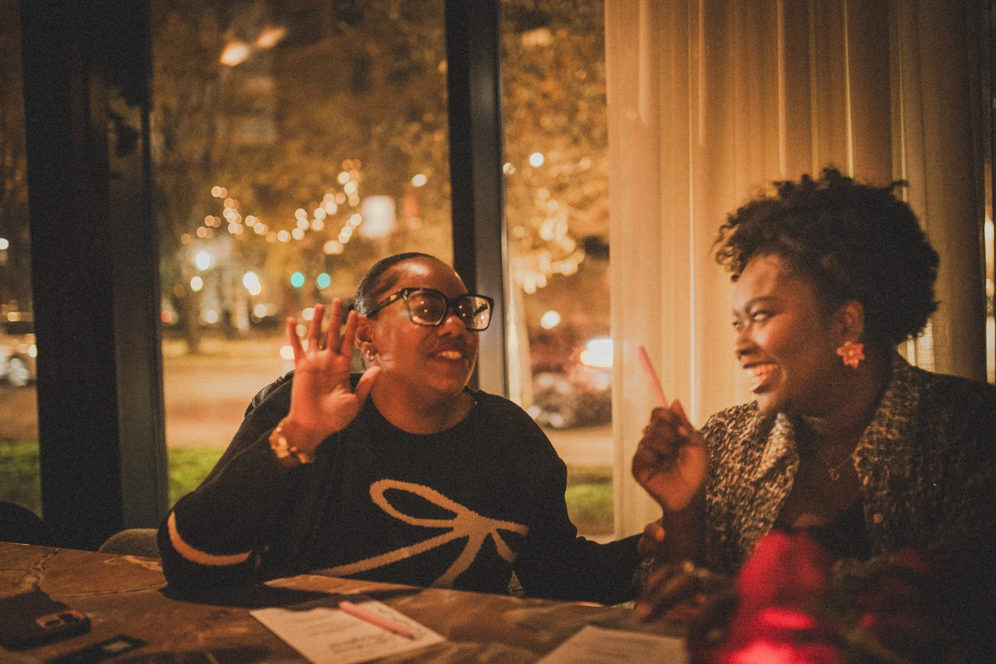 Two women are sitting at a table near a window, smiling and engaging in conversation. One woman, wearing glasses, has her hand raised to her ear. The other woman has a big smile and is gesturing with her hand. The scene appears to be indoors at night