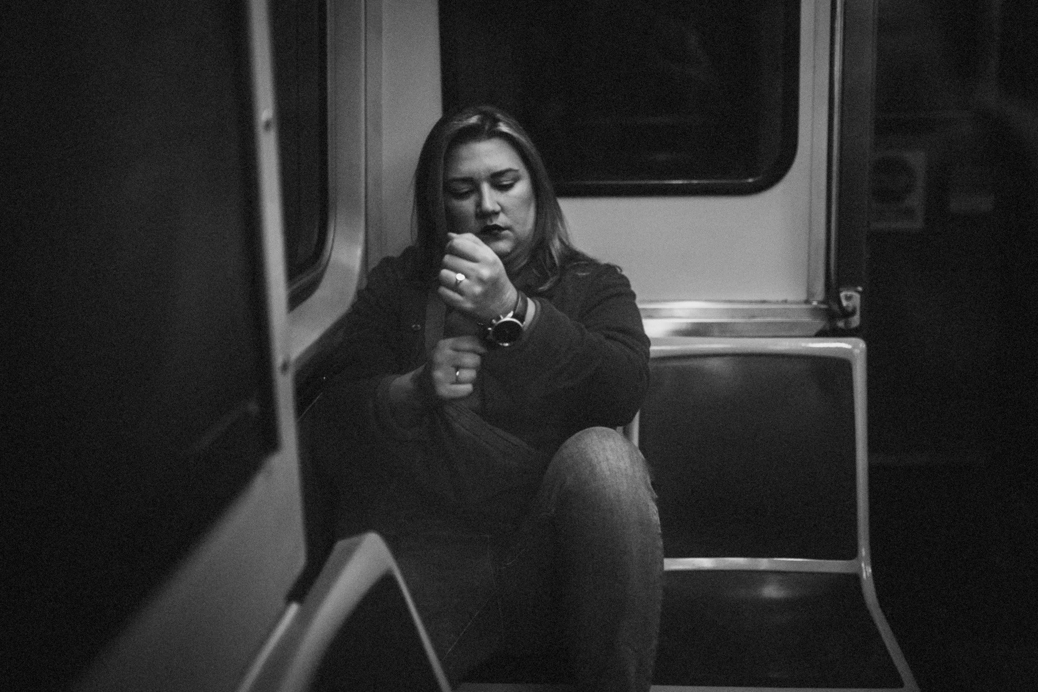 A woman with dark hair and dark clothing sits alone on a subway train, looking down and adjusting something in her hand, with a serious expression. She is wearing a watch and rings, and the image is in black and white.