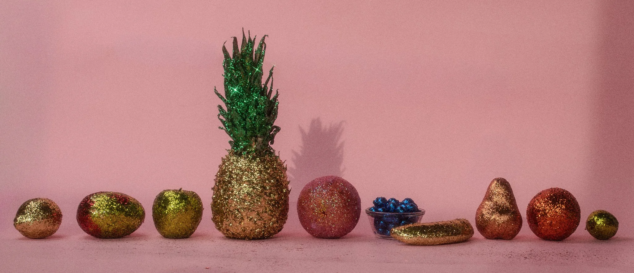 Decorative arrangement of fruits and objects covered in gold and glitter, including a pineapple, apples, a pomegranate, a bowl of berries, an avocado, and other items, all set against a pink background.