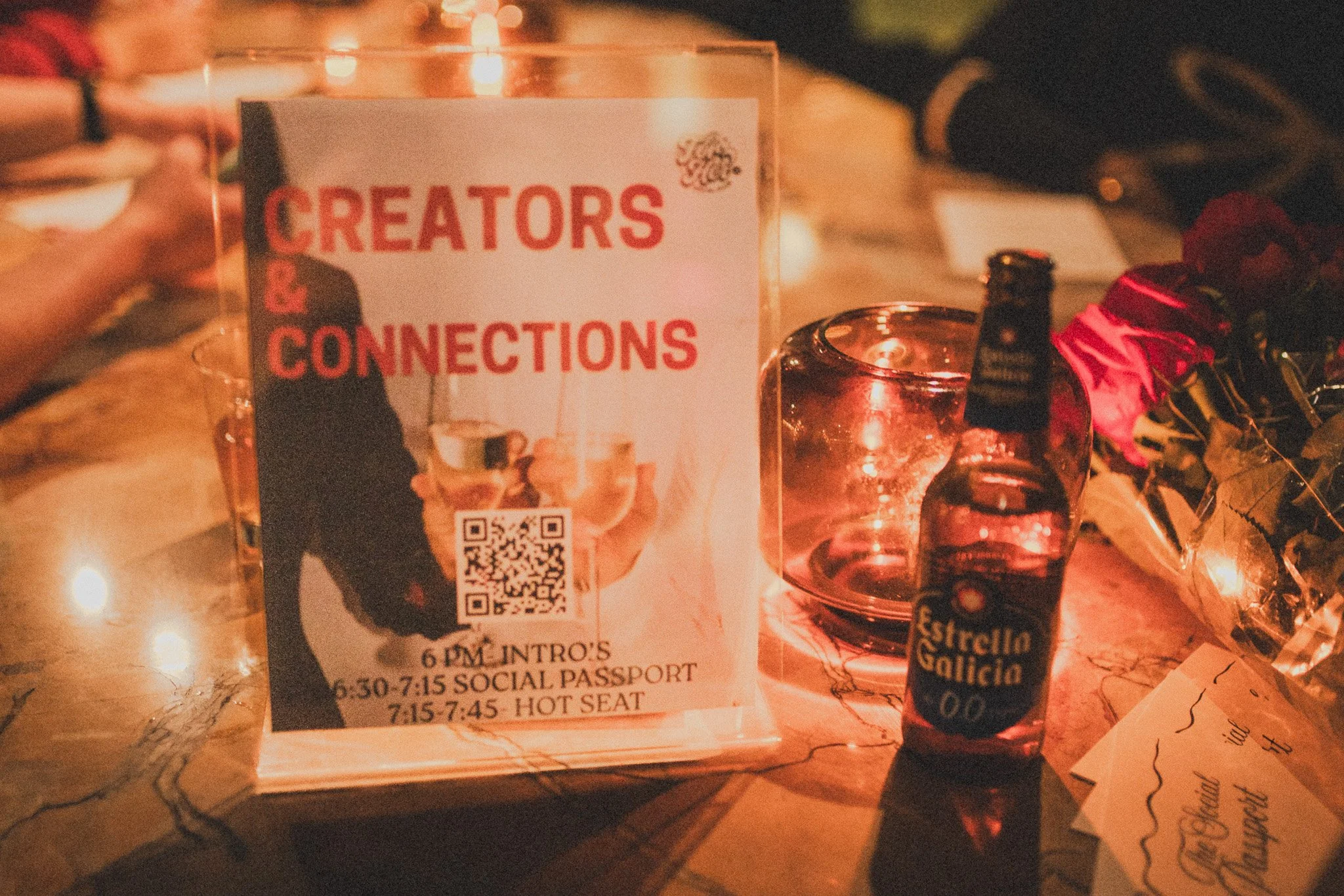 A table with a sign that reads 'Creators & Connections' with a QR code, a small bottle of Estrella Galicia beer, an empty glass, and some flowers in the background, illuminated by warm lighting.