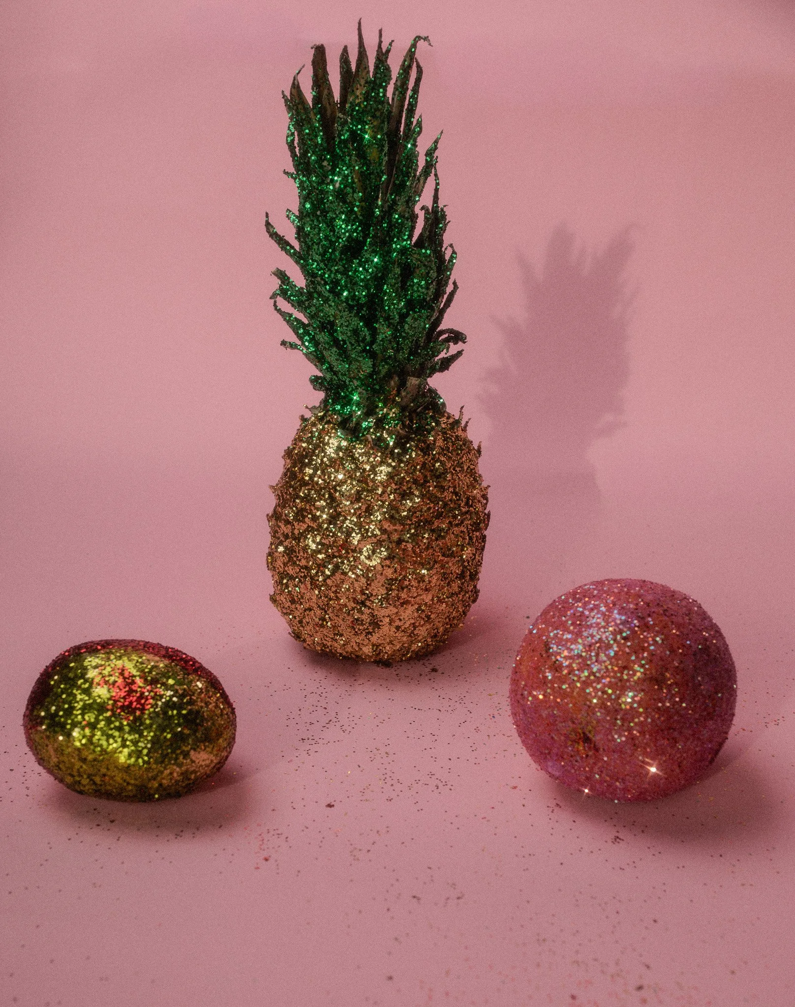 Decorative glitter pineapple standing between two glittery spheres on a pink background.