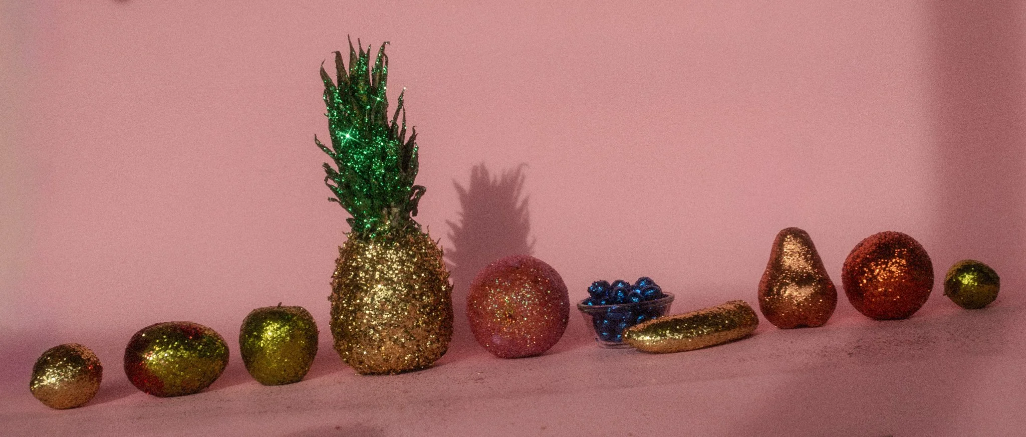 Decorative objects covered in gold glitter, including a pineapple with green glitter leaves, a red glitter ball, a small blue bowl with shiny berries, a gold glitter banana, and a gold and red glitter pear on a pink background.