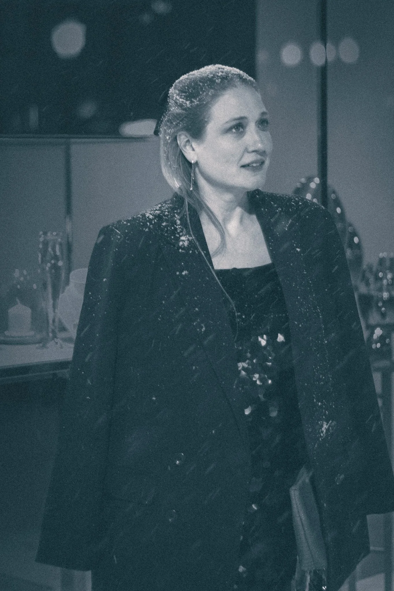 A woman with blonde hair styled in a bob, wearing a dark blazer and earrings, is looking slightly to her right. She is in a room with a mirror and various bottles and decorative items in the background.