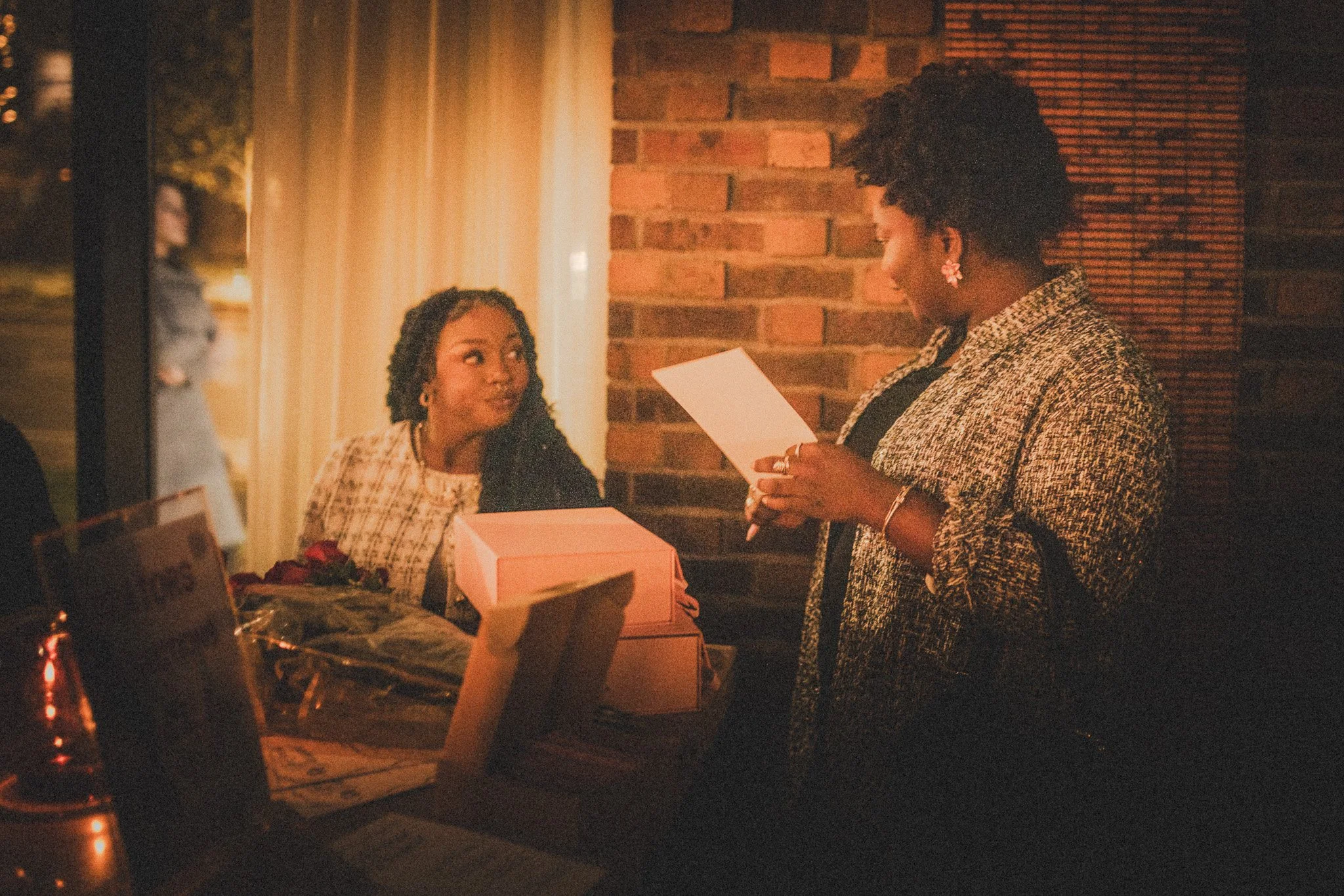 Two women at a table during an evening, one reading a card and the other looking surprised, with a bouquet of flowers and gift boxes nearby, against a brick wall background.