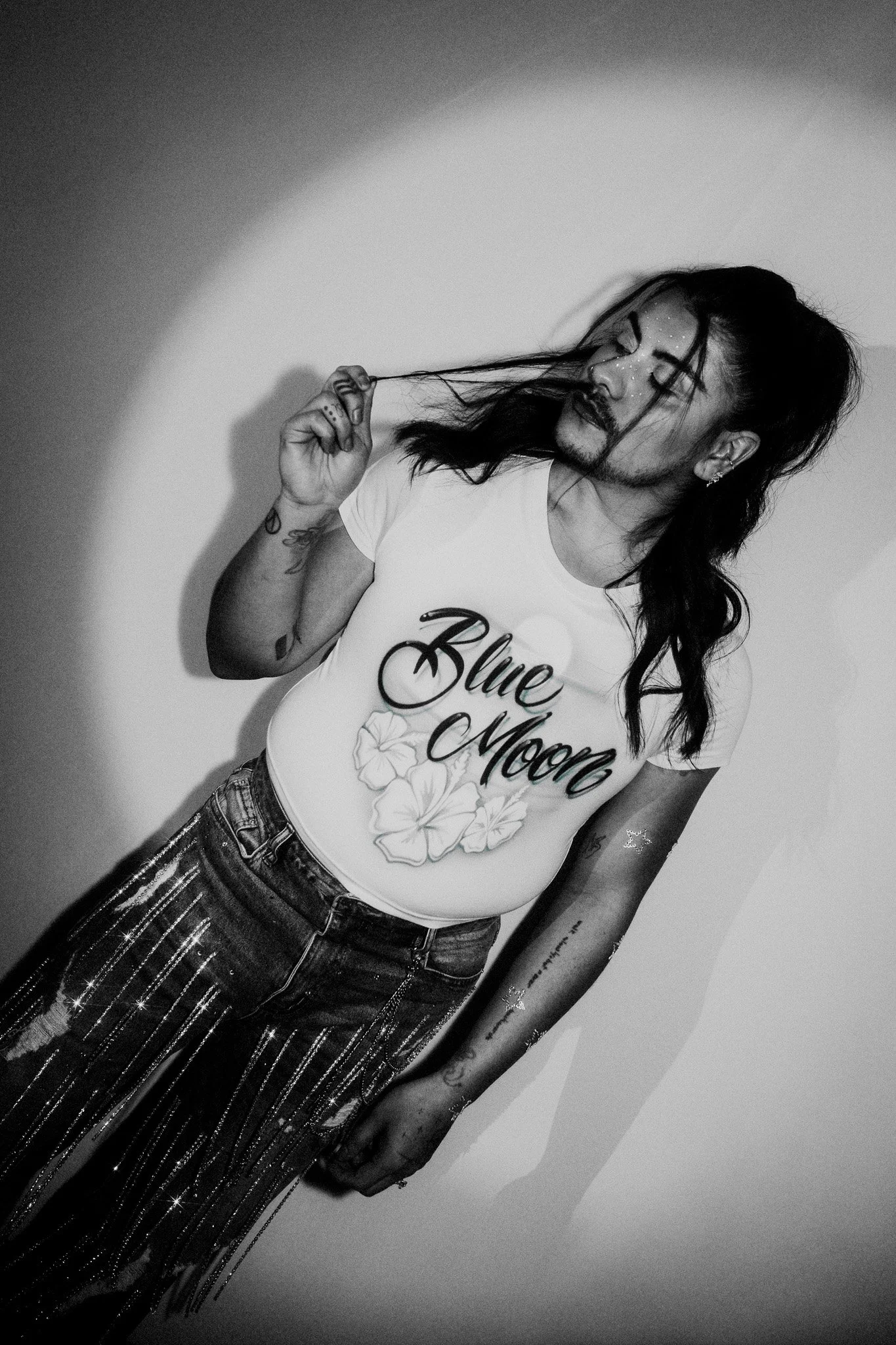 A person with long dark hair, tattoos, and dark lipstick poses with their eyes closed, wearing a white shirt with 'Blue Moon' and floral graphics, and sparkly fringed pants.