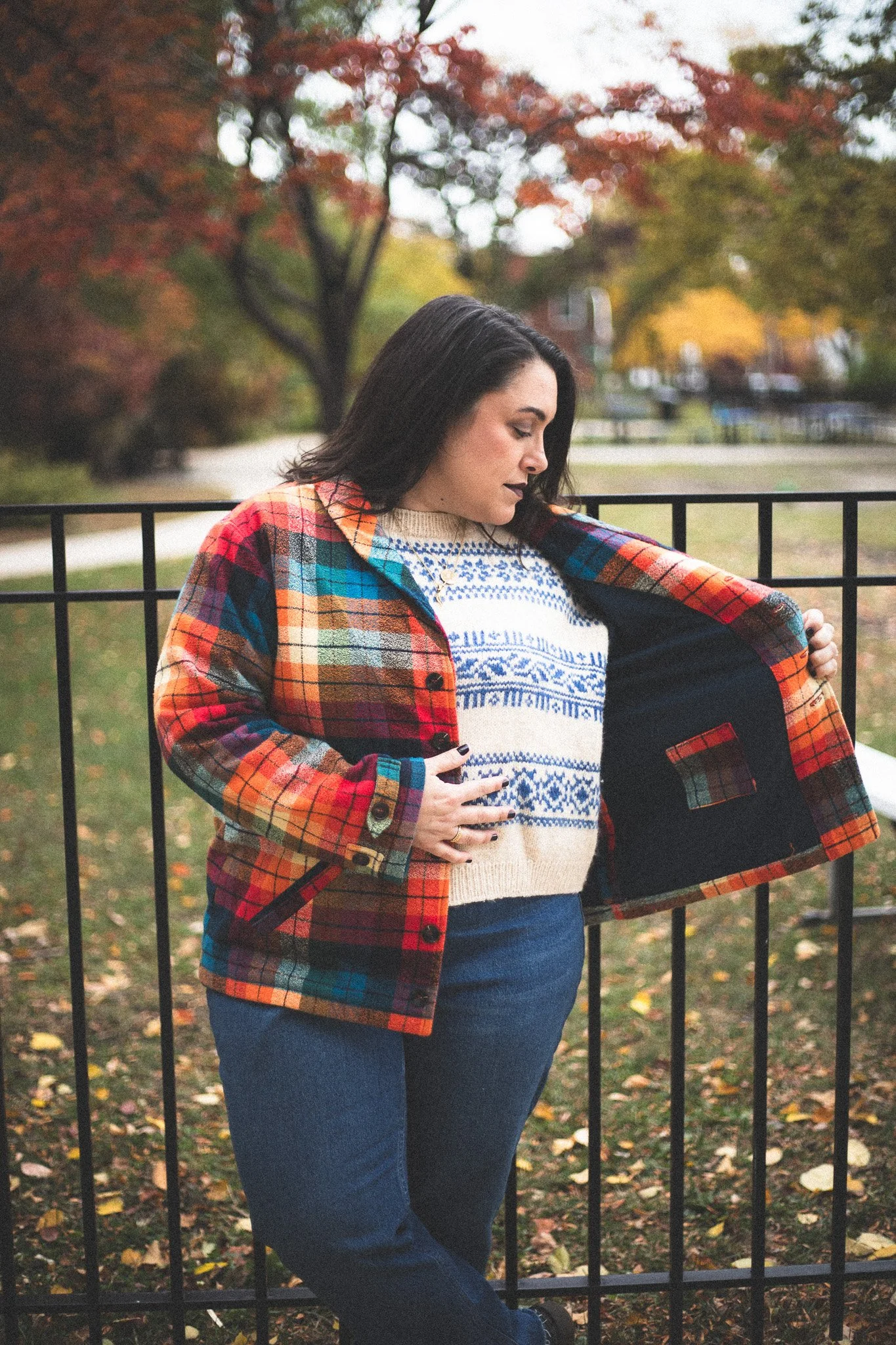 A woman in a colorful plaid jacket and patterned sweater standing outdoors by a black metal fence, with autumn leaves and trees in the background.