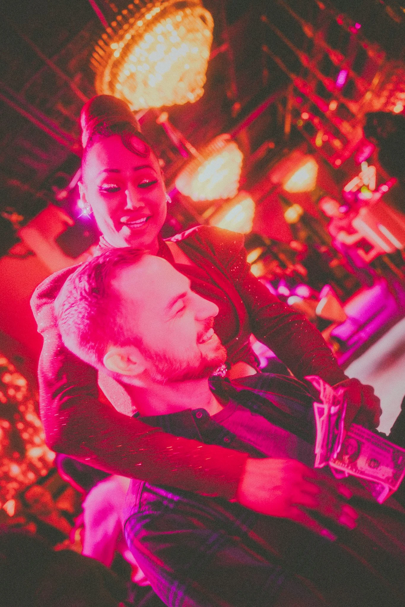 A woman with dark hair styled in vintage curls smiling and leaning over a man with light brown hair and a beard, who is also smiling. They are in a lively, colorful setting with bright decorative lights, possibly a nightclub or event venue.