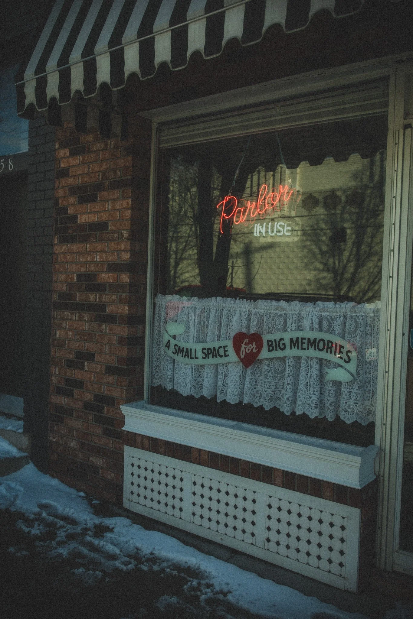 View of a storefront window with a neon sign that reads 'Parlor in Use' and a ribbon banner that says 'A small space for big memories', with lace curtains and a brick building exterior, during winter.