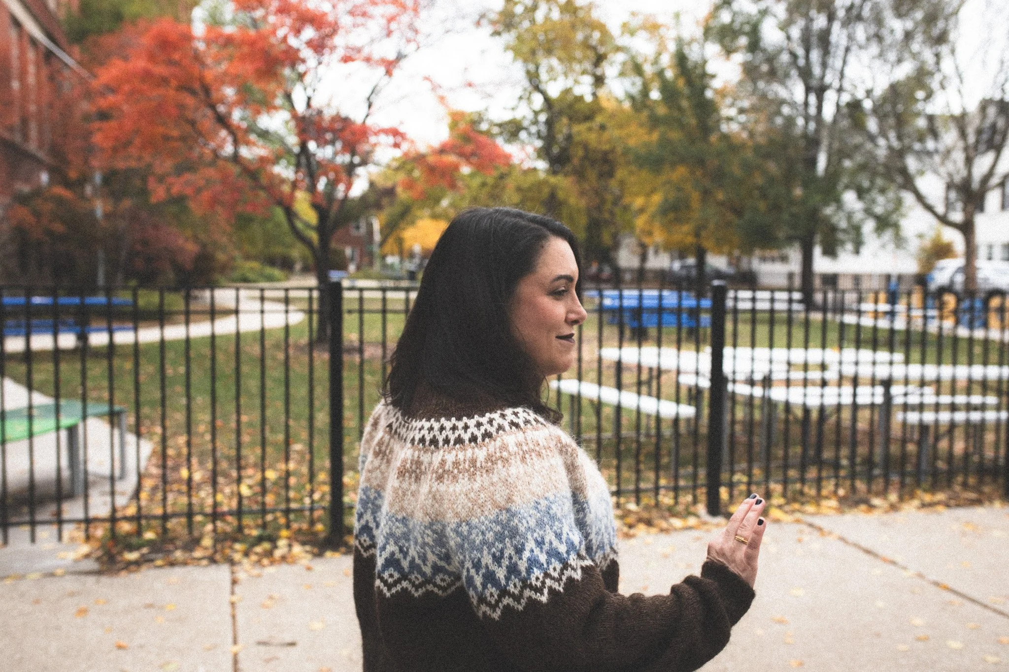A woman with dark hair wearing a patterned sweater standing outside in front of a black fence, with colorful autumn trees and a white building in the background.