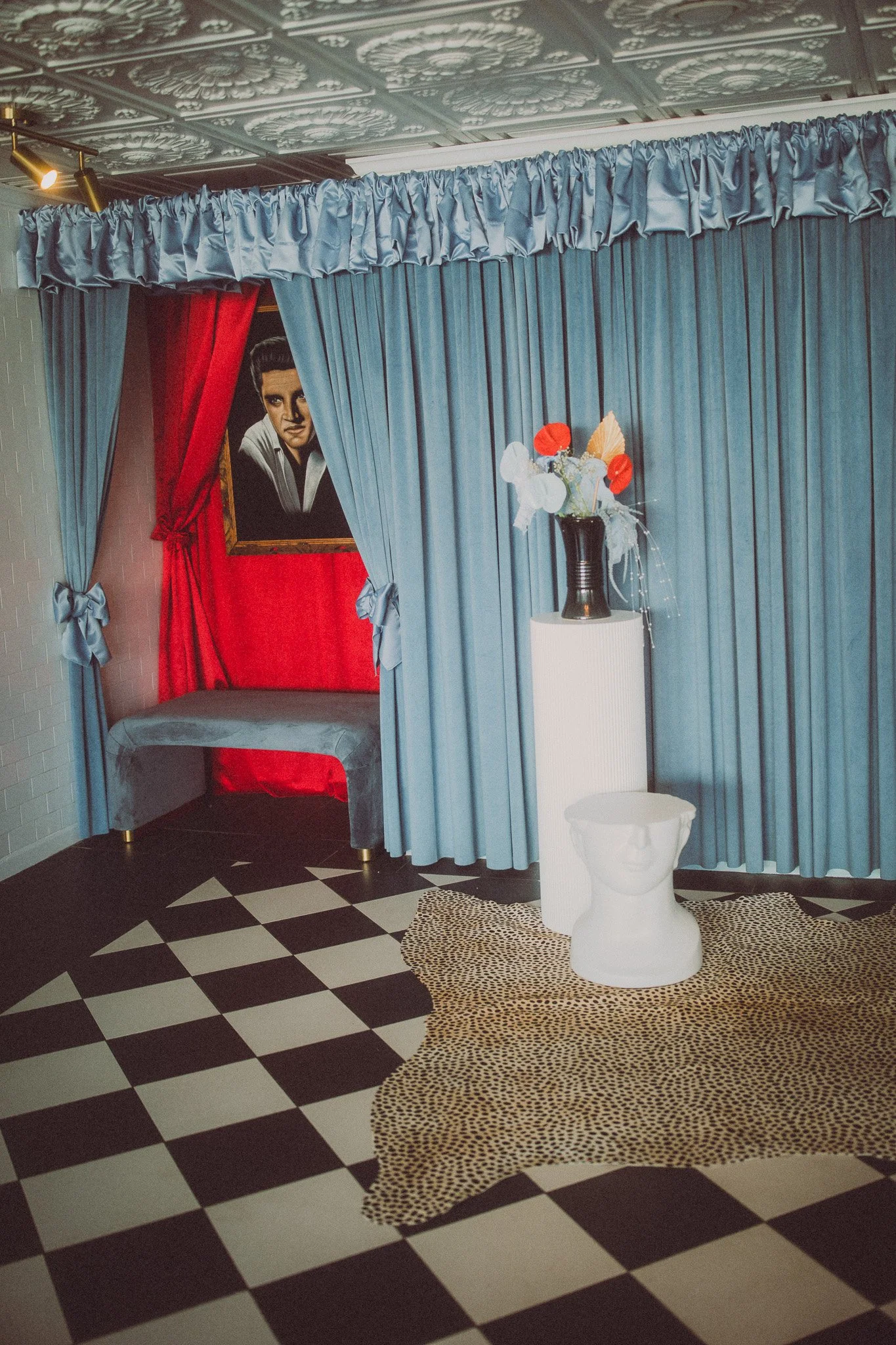 Interior with blue curtains, red and gray seating area, portrait painting of a woman, black vase with white, red, and orange flowers, white sculpture of a face on leopard-print rug, black and white checkered floor.