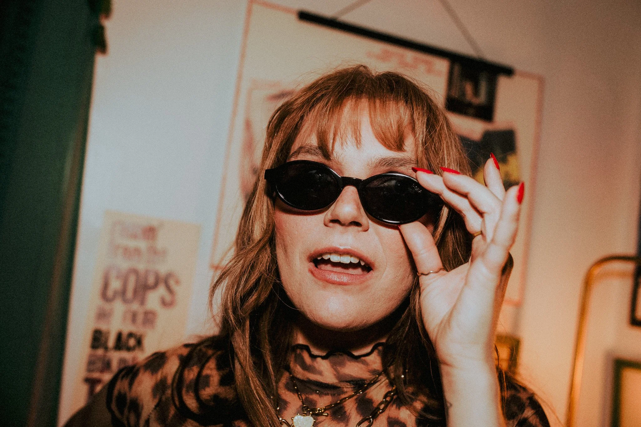A woman with brown hair and bangs adjusting her black sunglasses with her right hand, wearing a leopard print top, necklaces, and red nail polish, in a room with posters on the wall.